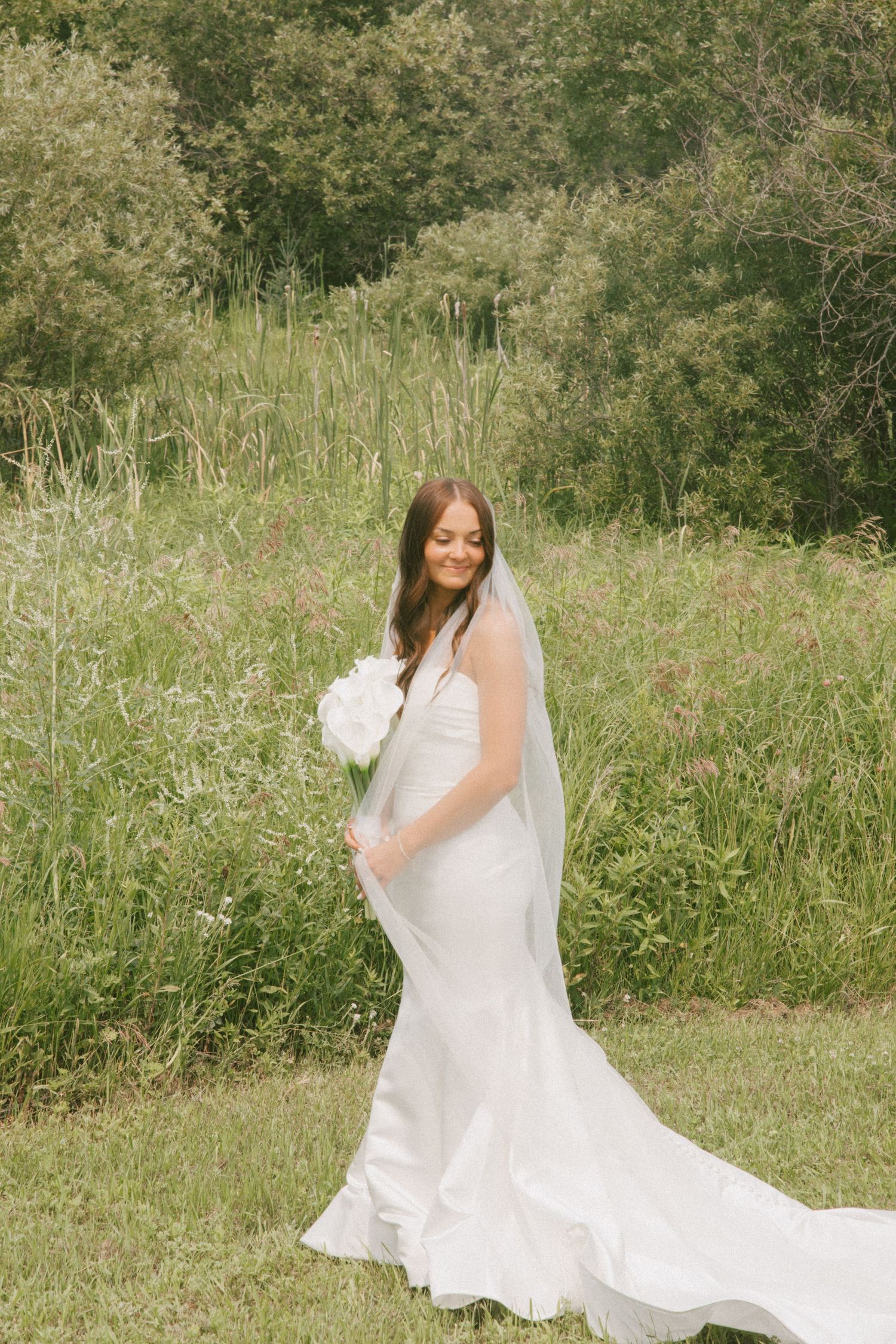 Elegant bride in fitted white gown holding a white bouquet while standing in a wildflower meadow.
