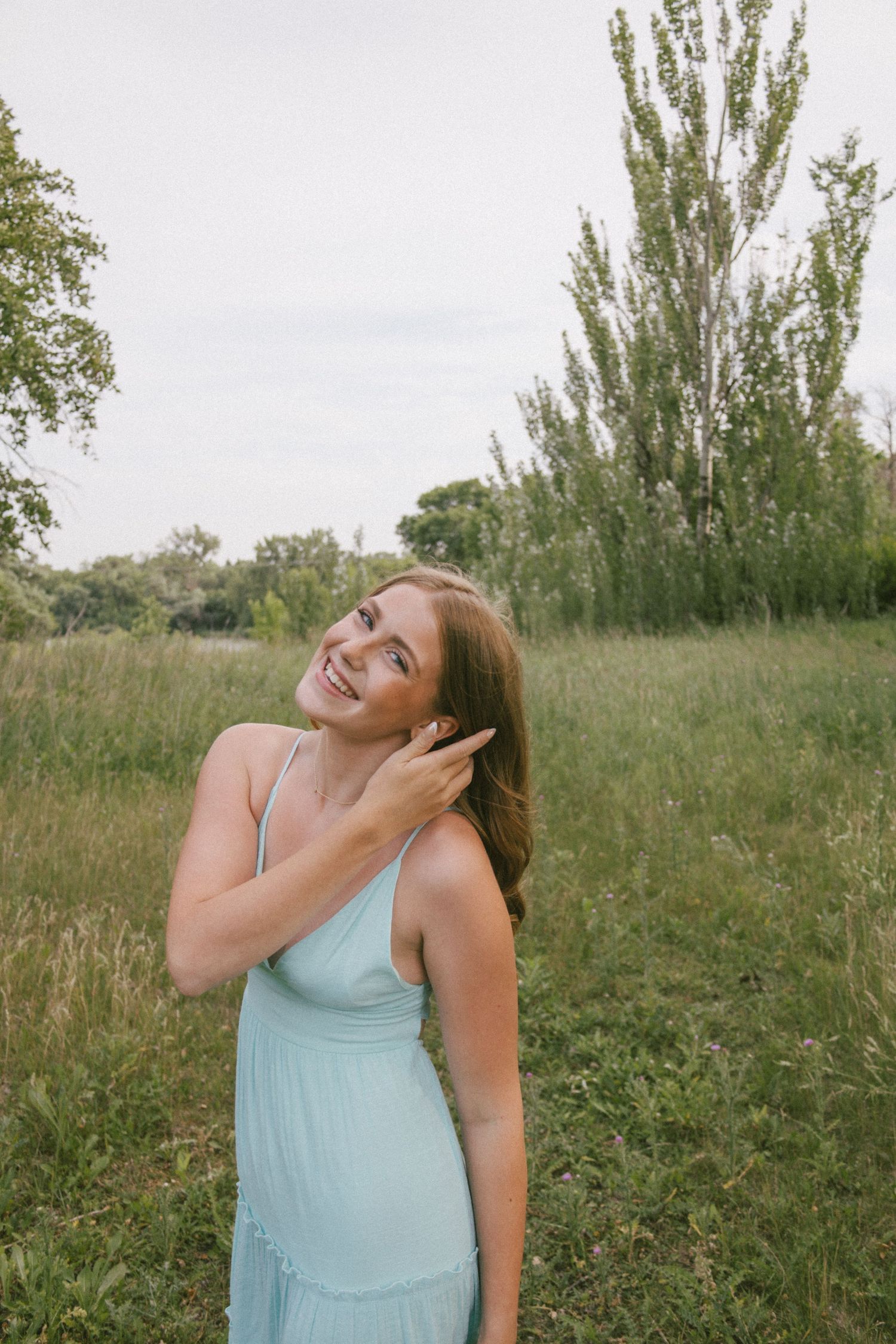 Young woman in mint green dress smiling outdoors in lush green field with trees in background.