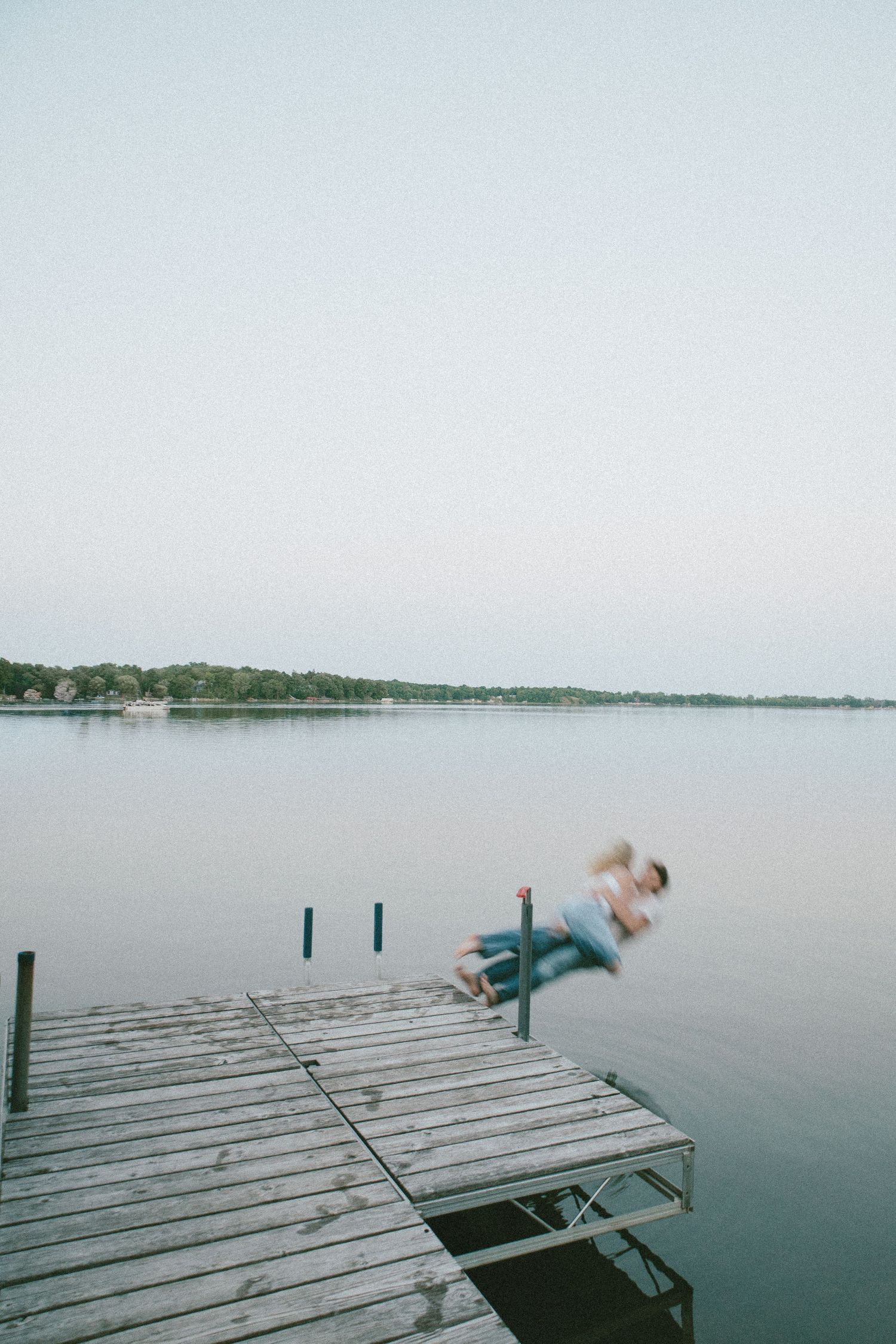 Person diving off a wooden dock into a calm lake at dusk with a tree-lined shore in the background.