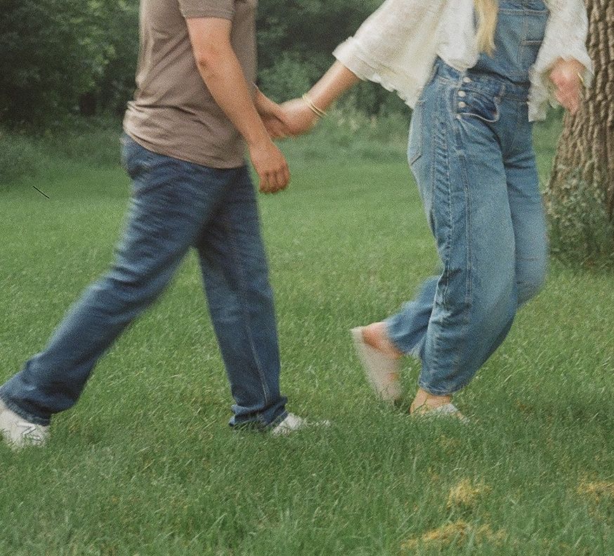 Couple holding hands walking on green grass, wearing jeans and casual shoes in an outdoor park setting.