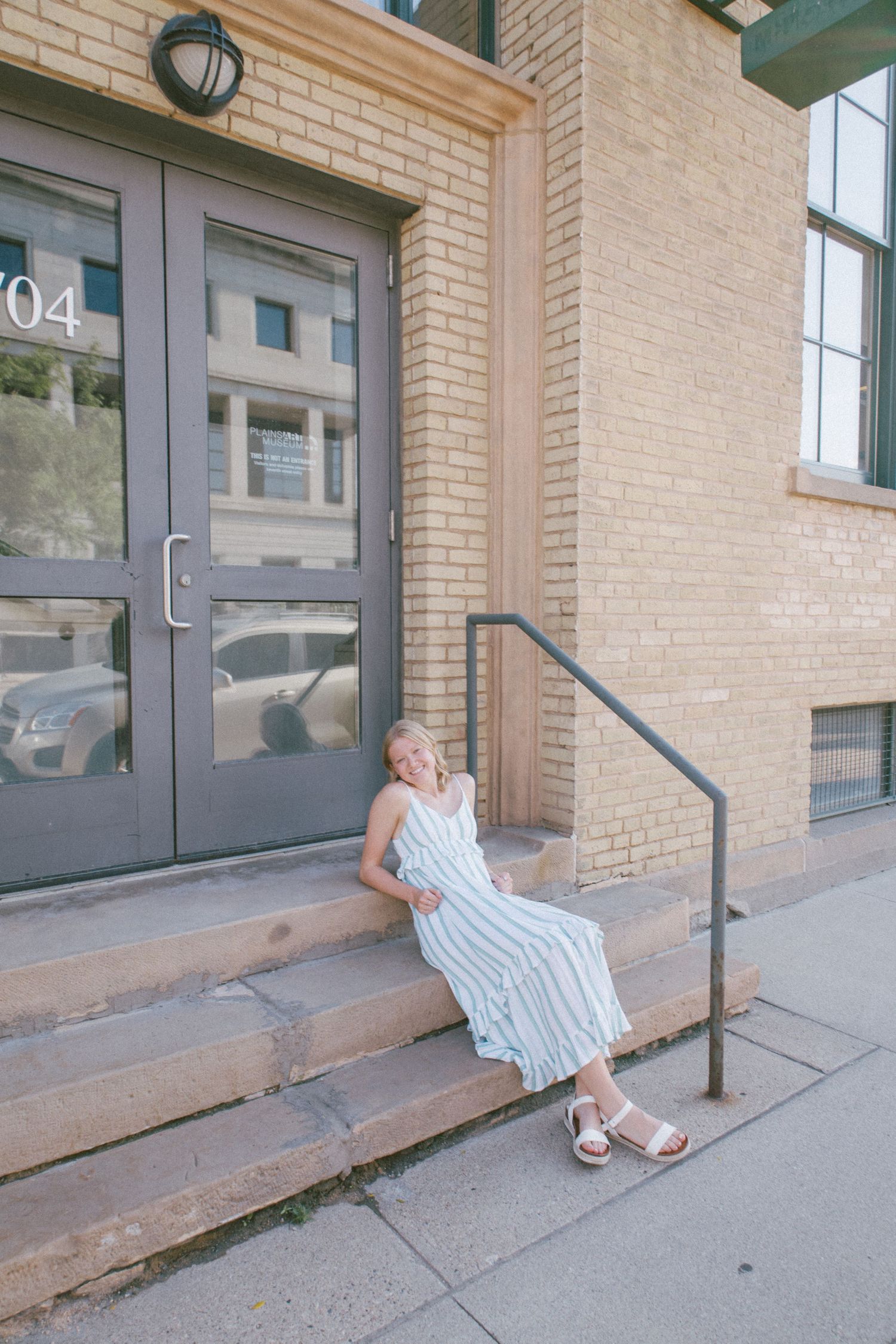 Woman in striped midi dress sitting on brick building steps, smiling outdoors in summer.