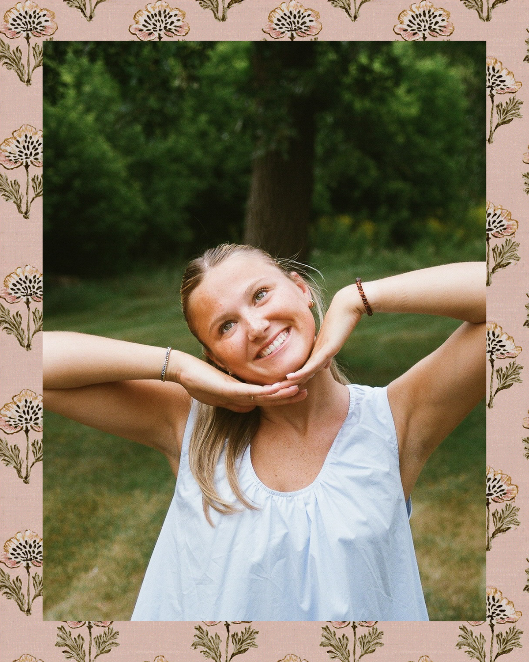 Smiling young woman in white dress posing outdoors with hands behind head, framed by floral pink border.