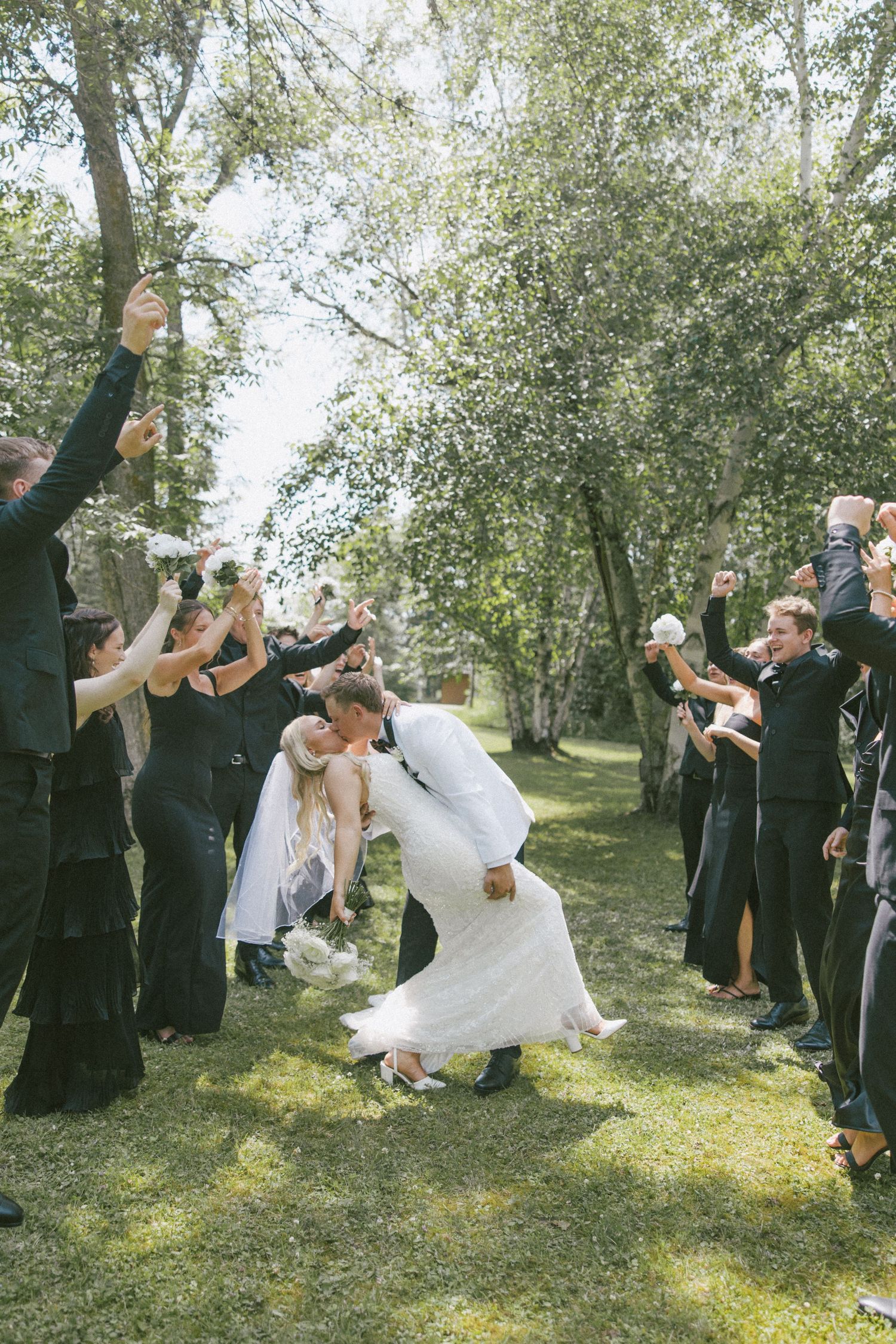 Bride and groom share a romantic dip kiss while wedding guests cheer in a tree-lined outdoor aisle.