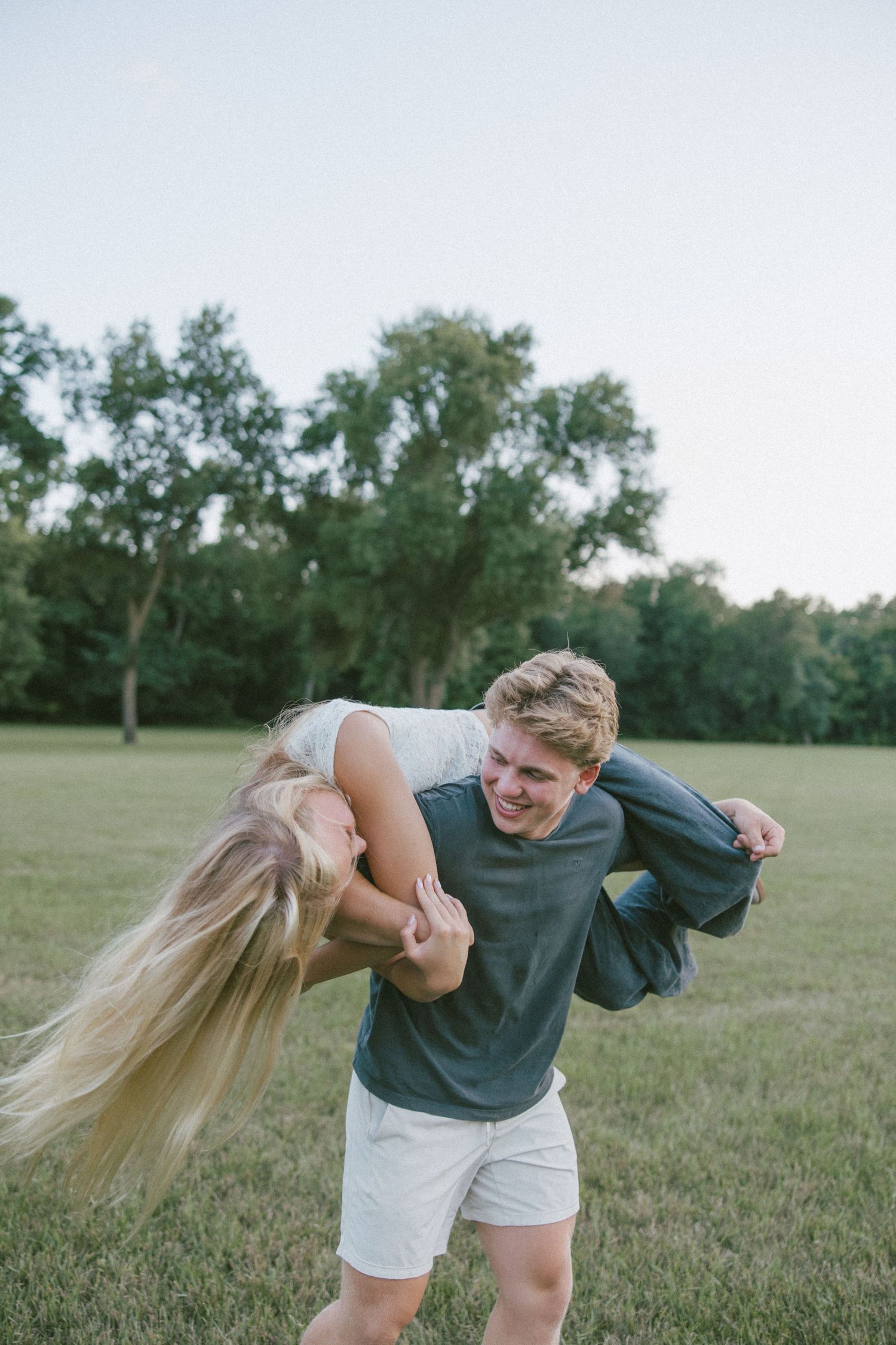 A young man carries a laughing blonde woman over his shoulder in a sunny green park.