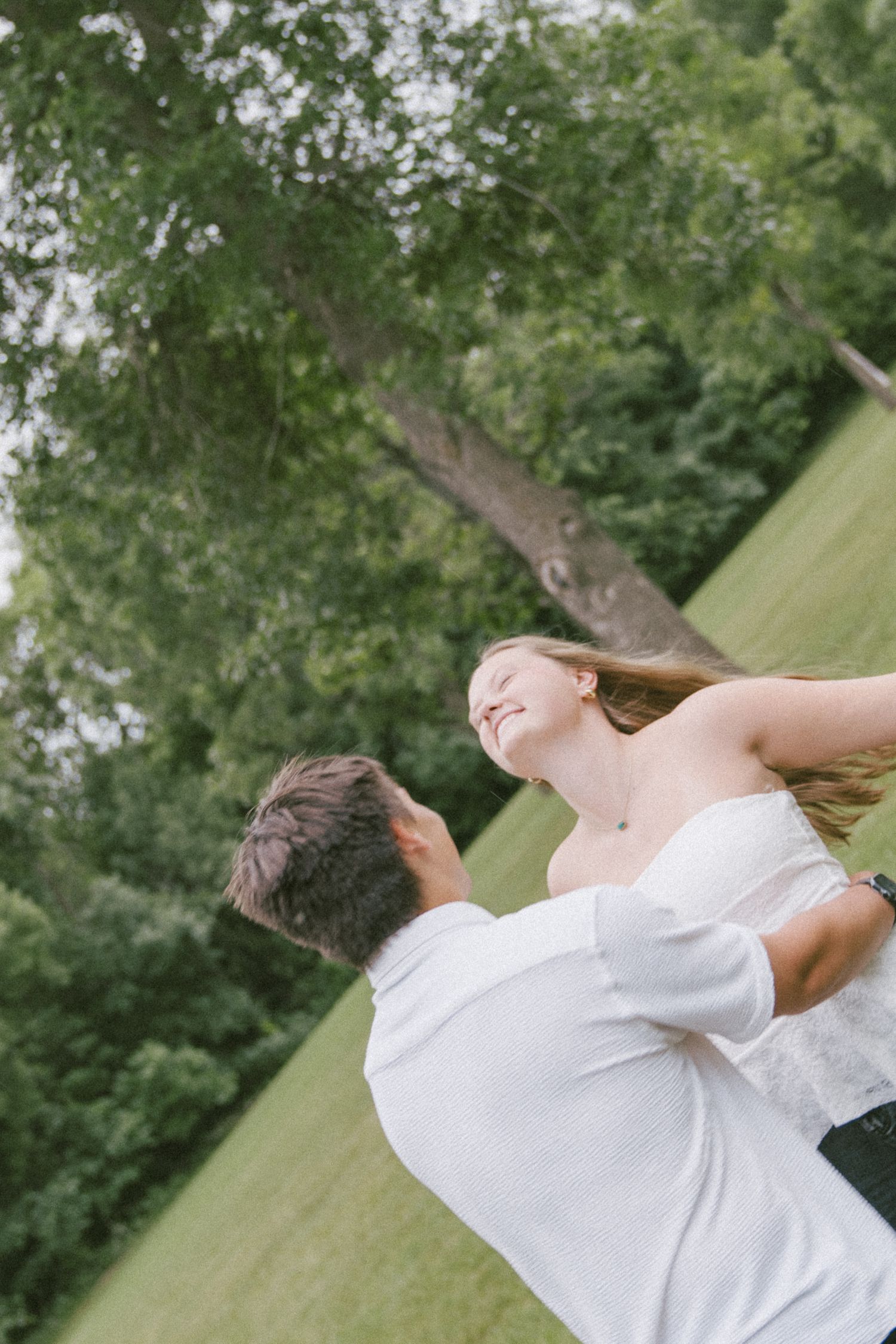 A groom lifts his smiling bride outdoors in a lush green park setting during a romantic wedding celebration.
