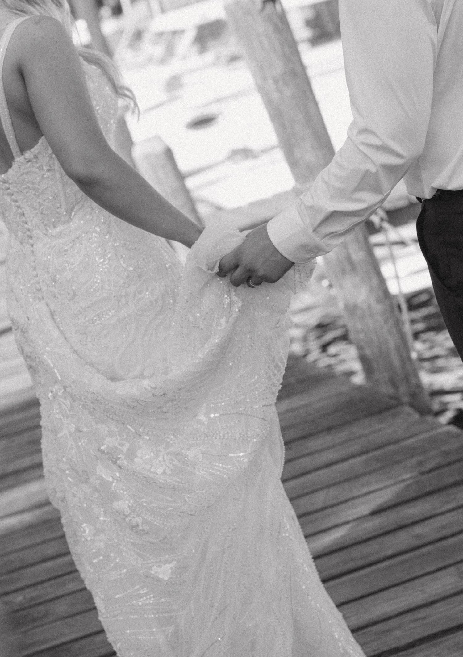 Black and white photo of a bride and groom holding hands on a wooden dock, bride wearing a beaded lace wedding gown.