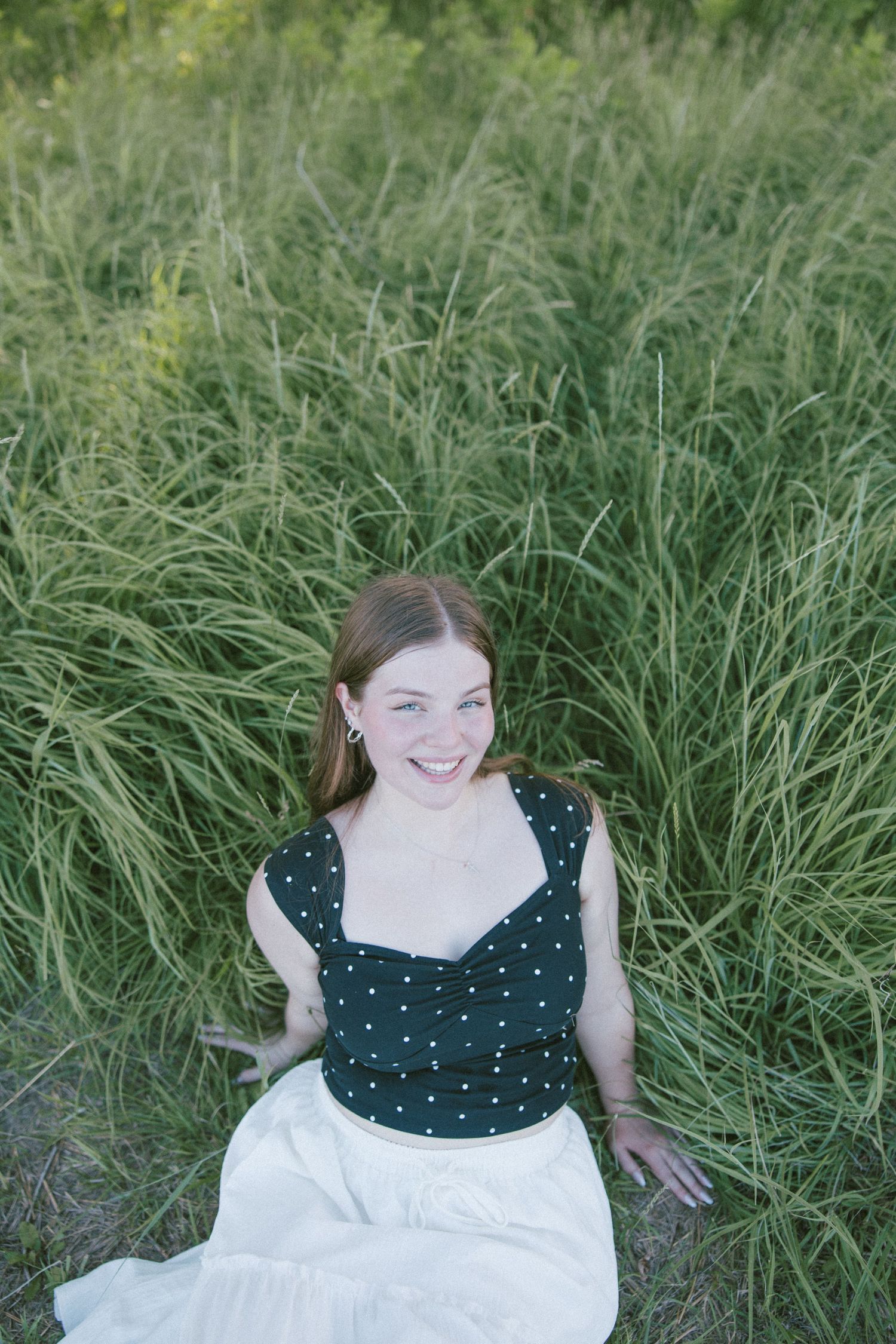 Smiling young woman in polka dot top and white skirt sitting in tall green grass outdoors.