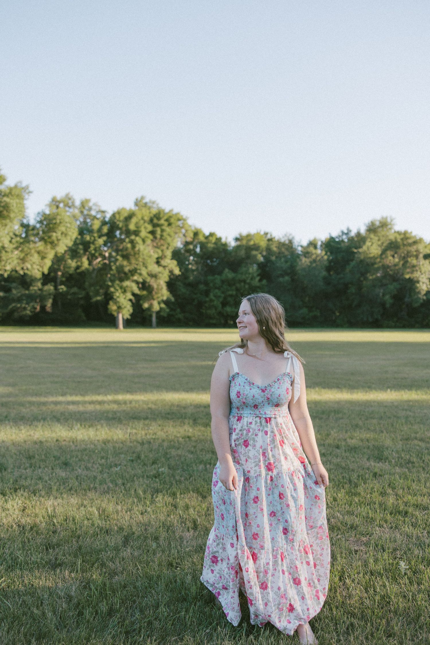 Young woman in floral maxi dress smiling in green field with trees in background on sunny day.