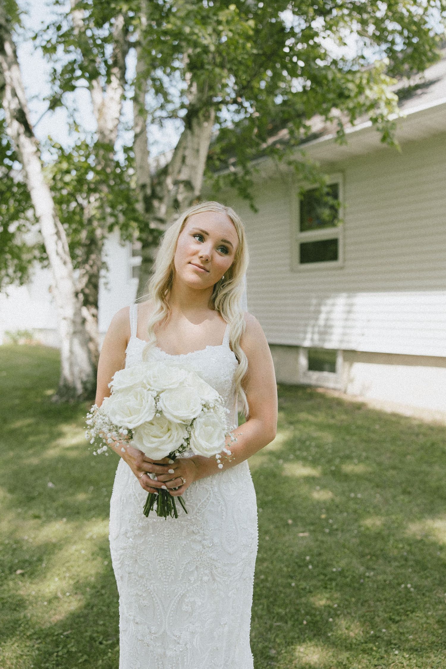 Blonde bride in white lace gown holds white rose bouquet outdoors near birch tree and white building.