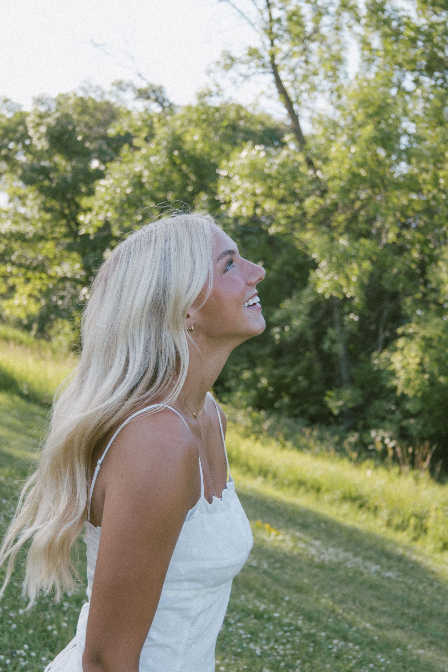 Smiling blonde woman in white sundress laughing outdoors in lush green park on sunny day.