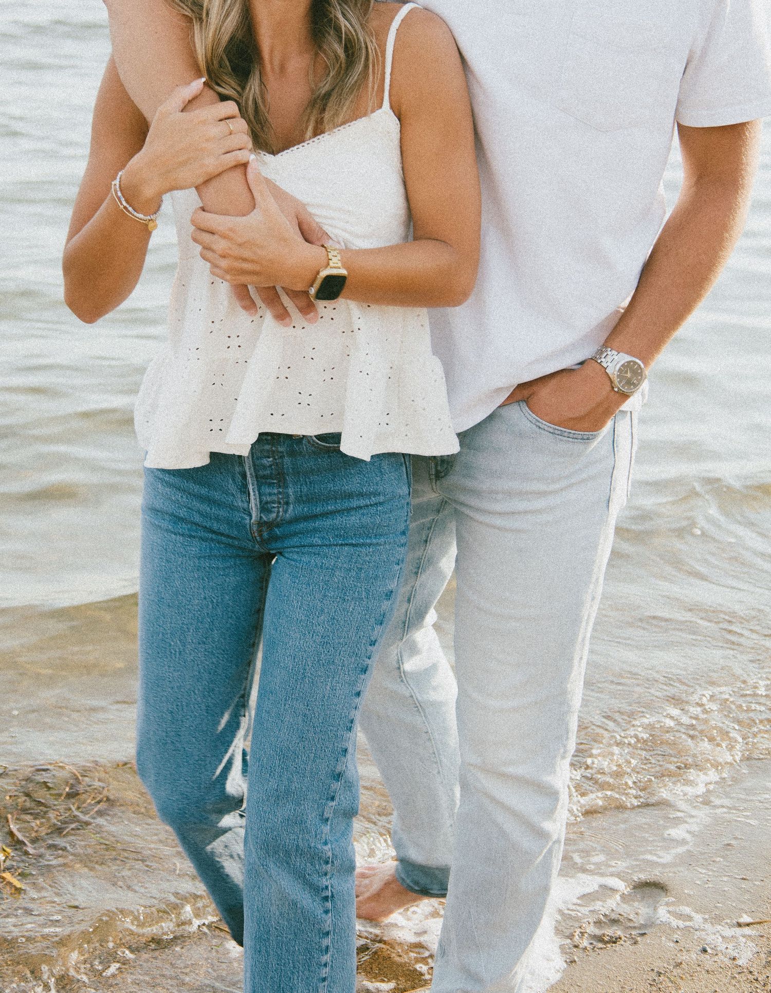 Couple embracing on a beach, wearing white tops and light blue jeans, standing in shallow water.