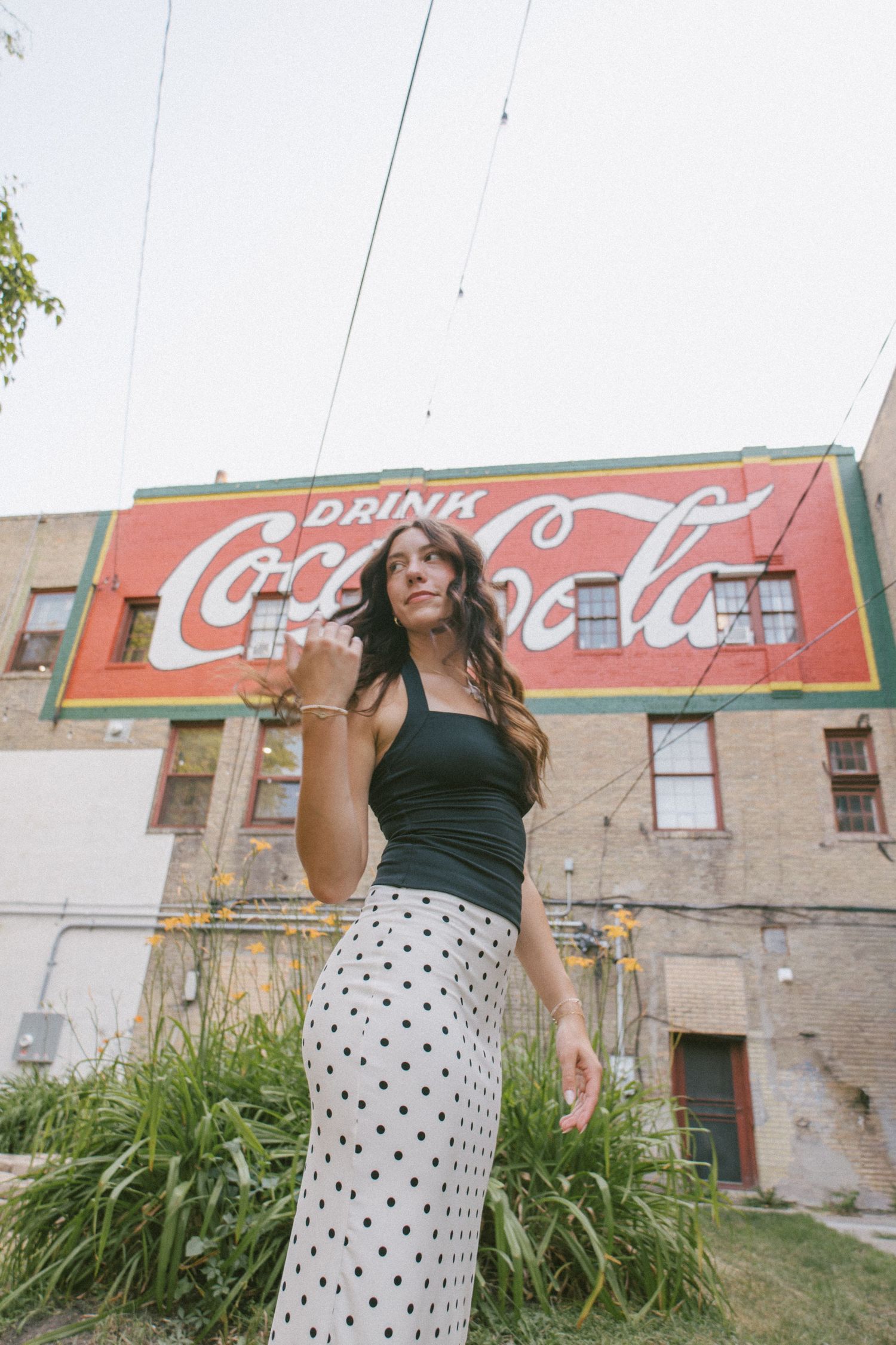 Woman in black top and polka dot skirt posing in front of a vintage Coca-Cola mural on a brick building.