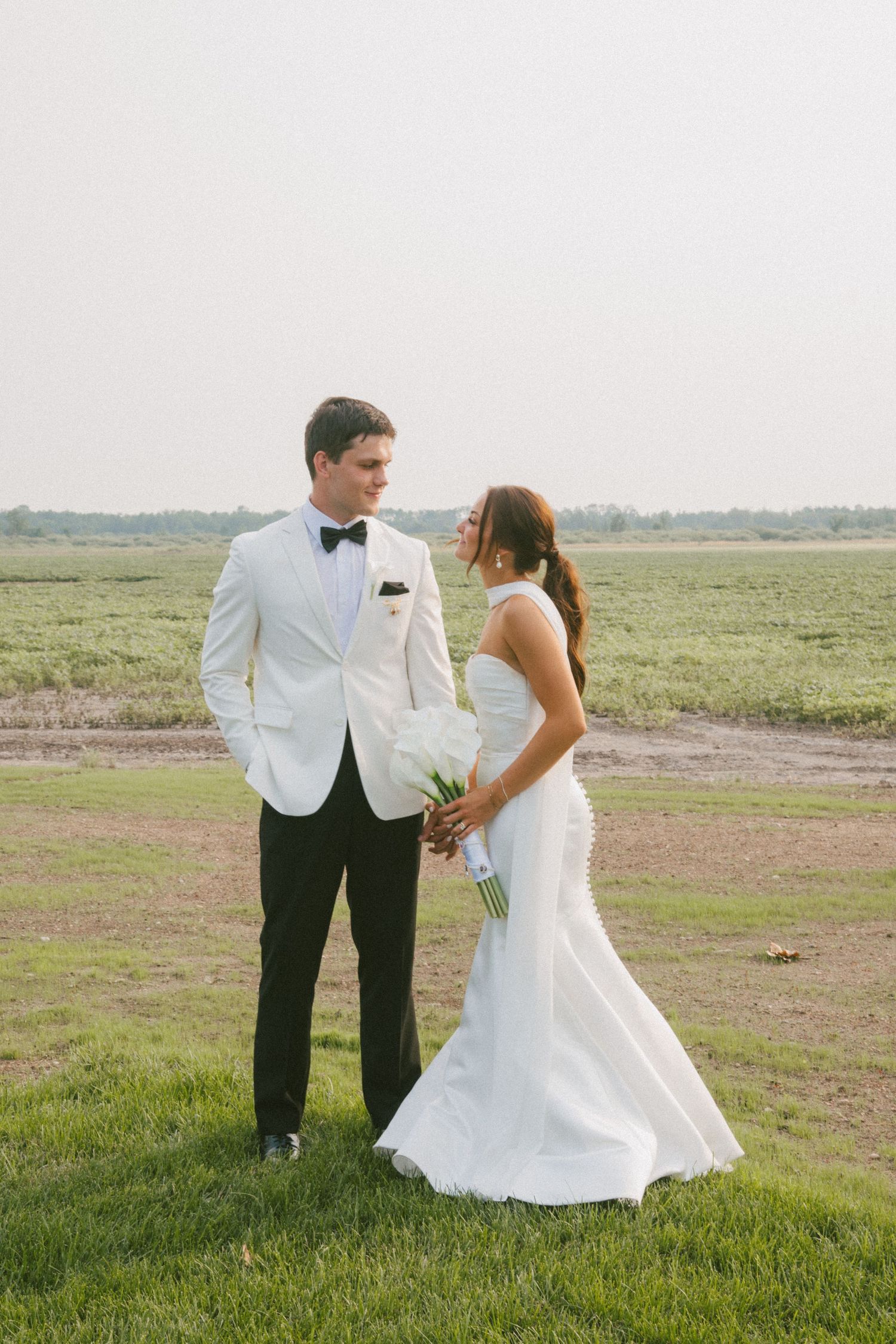 Bride and groom in elegant wedding attire sharing a tender moment overlooking a vast green field.