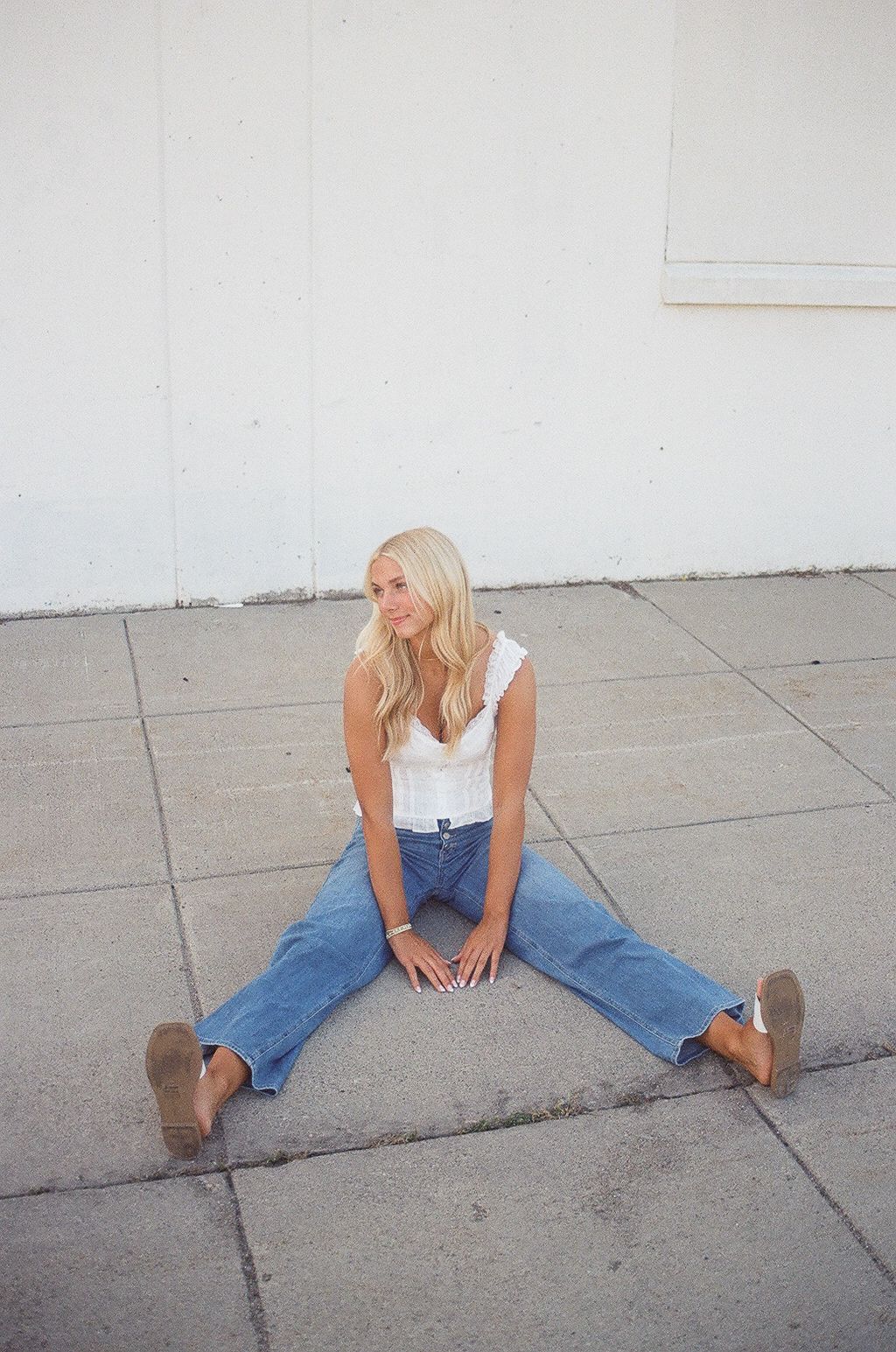 Blonde woman in white top and blue jeans sitting on concrete ground outdoors against a white wall.
