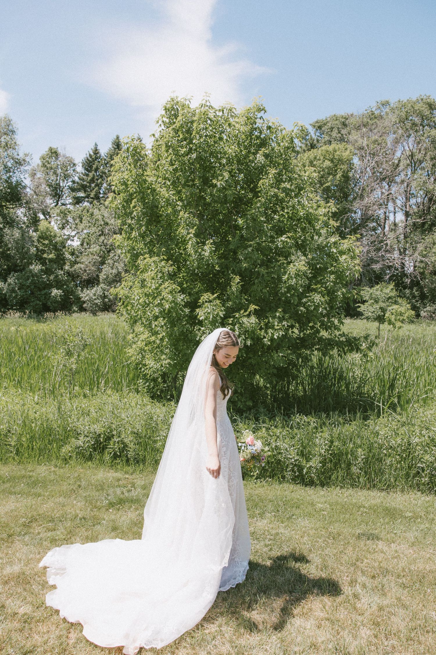 Bride in white gown and long veil stands gracefully on green grass with lush trees in background on sunny day.