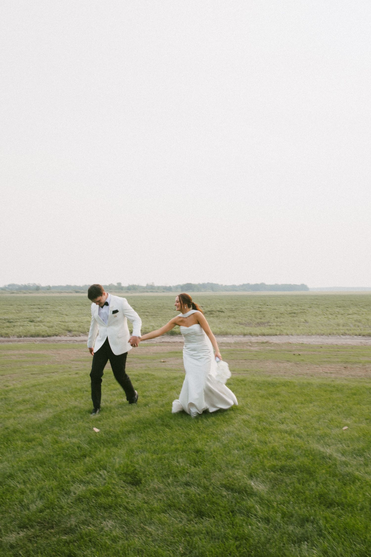 Bride and groom hold hands and dance playfully on a lush green field on their wedding day.
