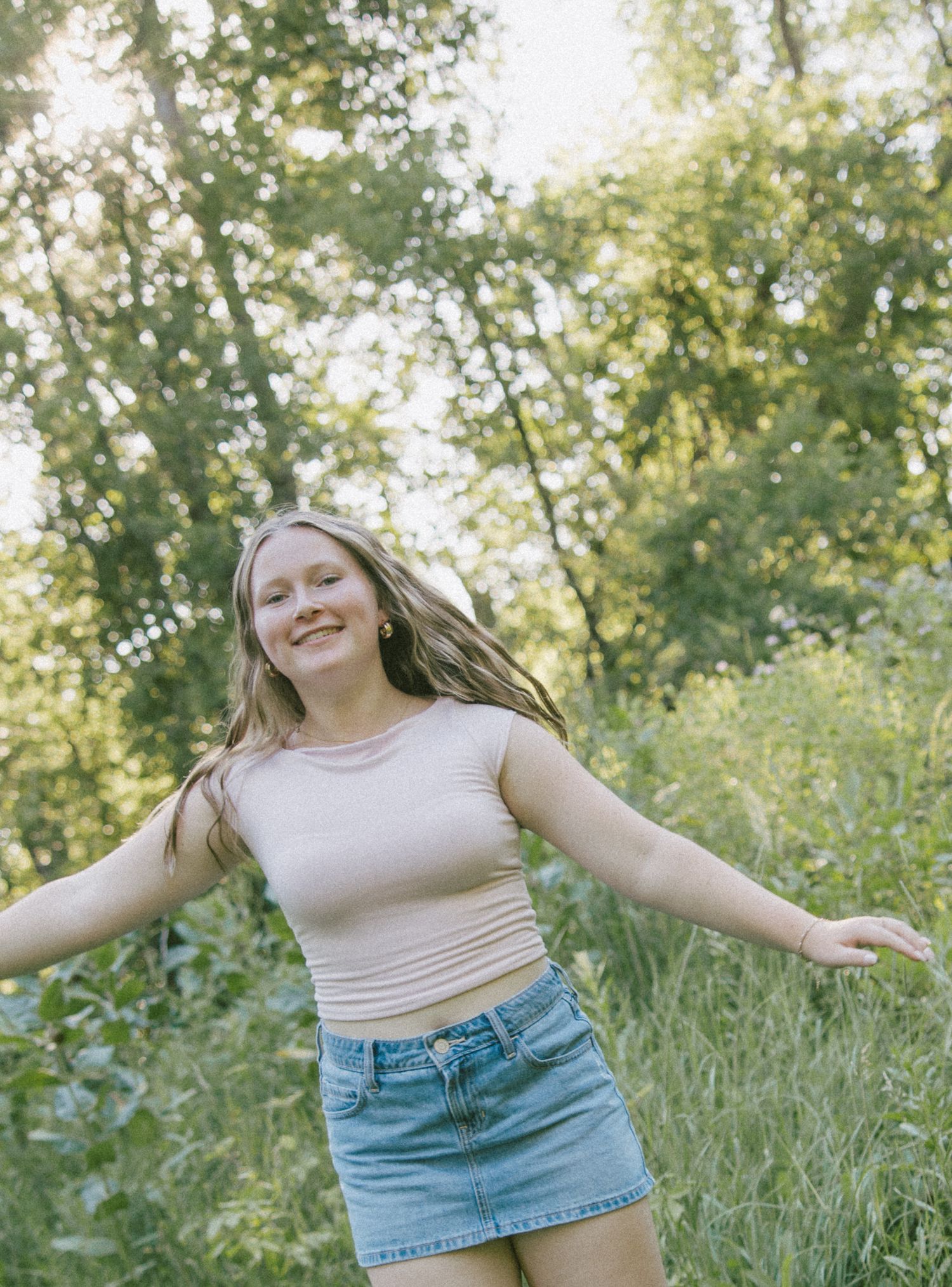 Young woman in crop top and denim skirt smiling with arms outstretched in a sunny green forest setting.