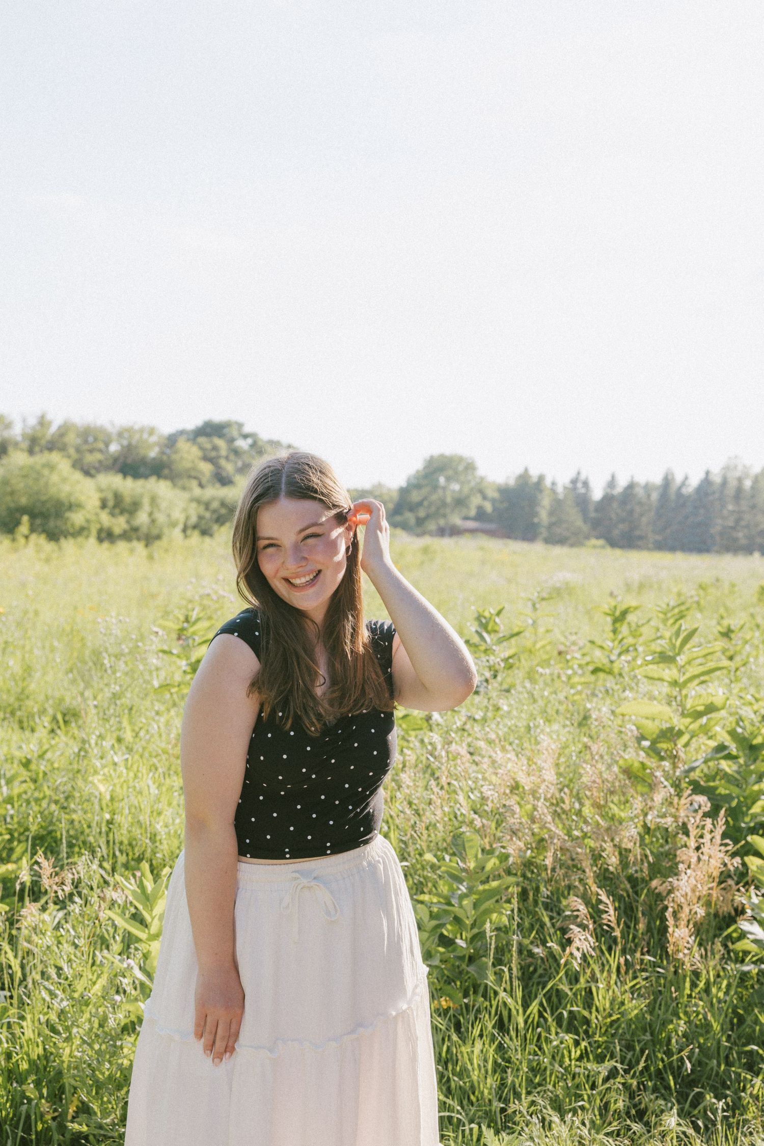 Smiling young woman in polka dot top and white skirt standing in a sunny green meadow.