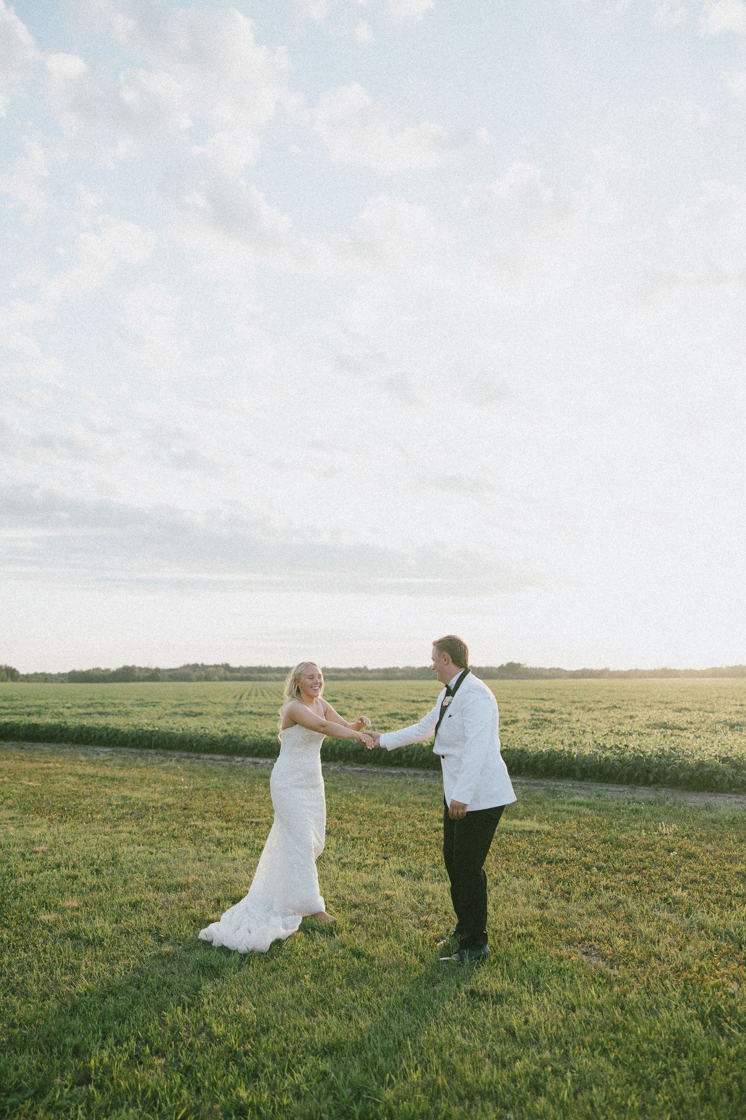 Bride and groom holding hands and dancing in a lush green field at golden hour on their wedding day.