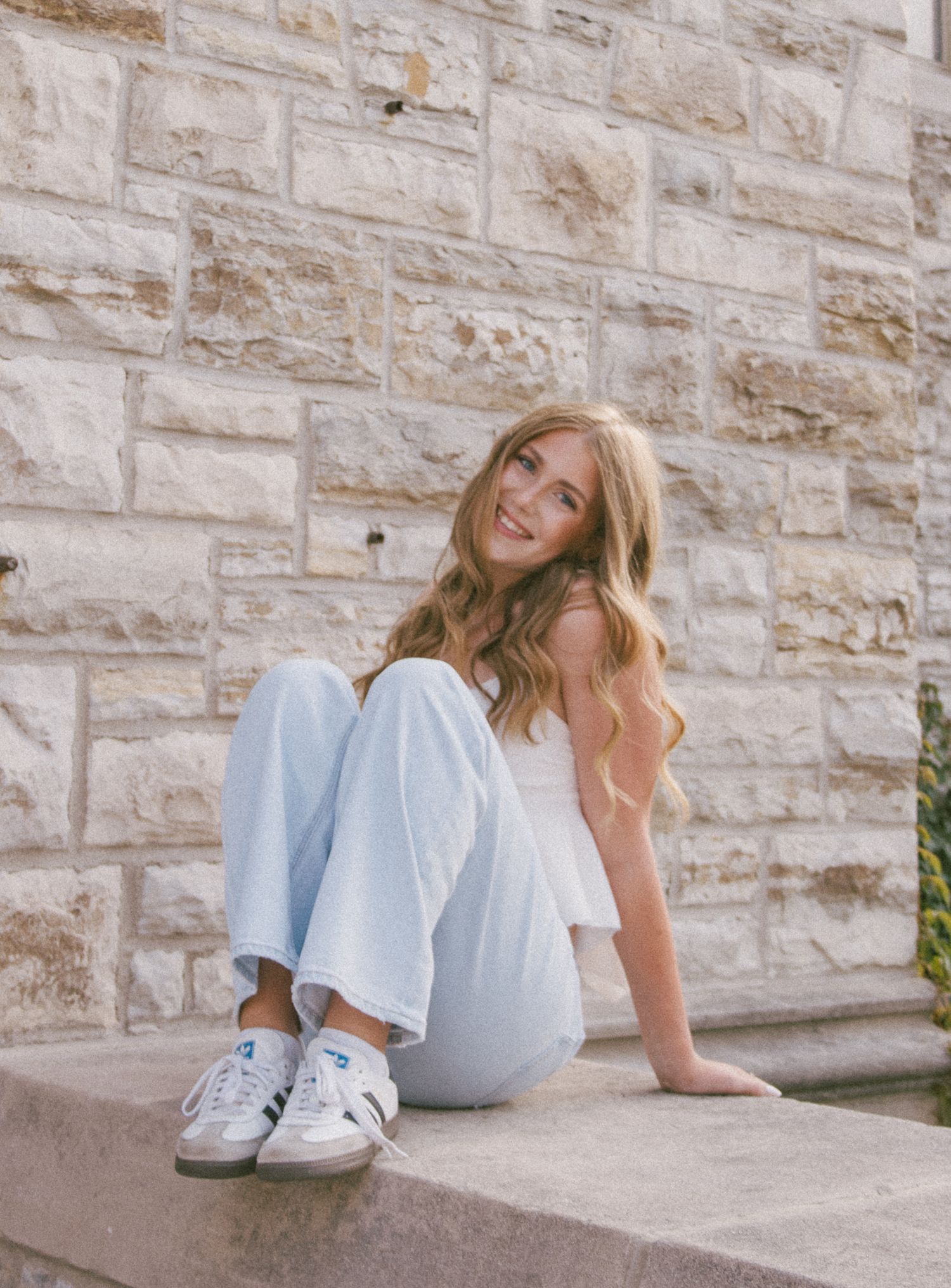 Smiling young woman with long blonde hair sitting on stone steps wearing white top, light blue wide-leg pants, and white sneakers.