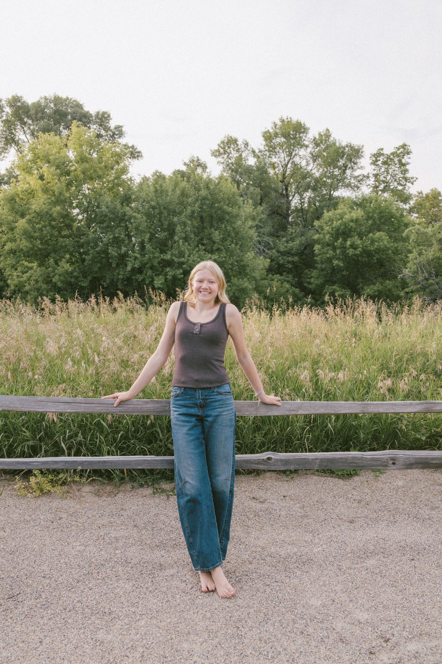 Smiling blonde woman in jeans and tank top leaning on wooden fence with green meadow and trees behind her.