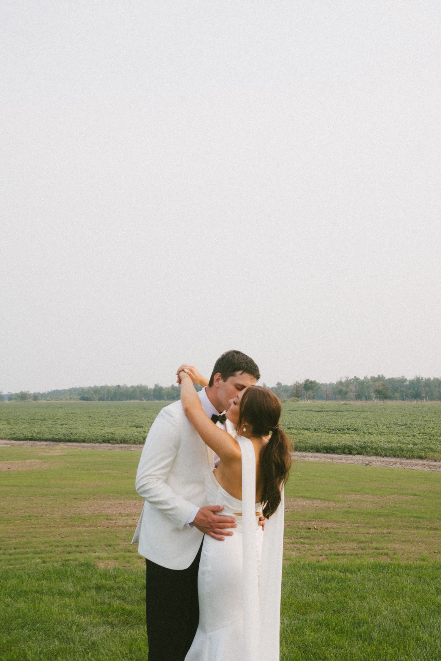 Groom in white tuxedo kisses bride in elegant backless white gown outdoors on green field.