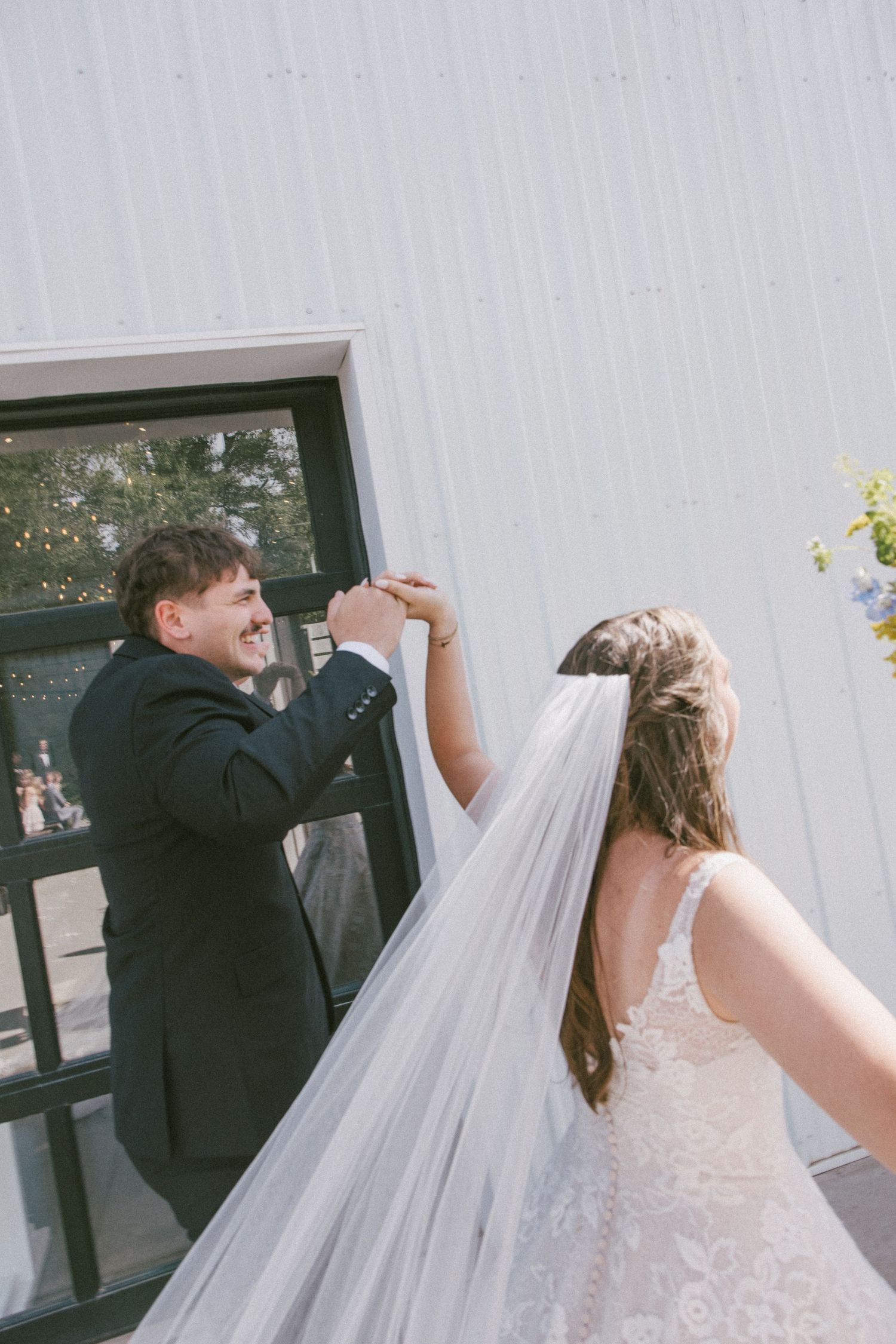 A groom in a black suit smiles while spinning his bride in a white lace gown and veil outdoors.