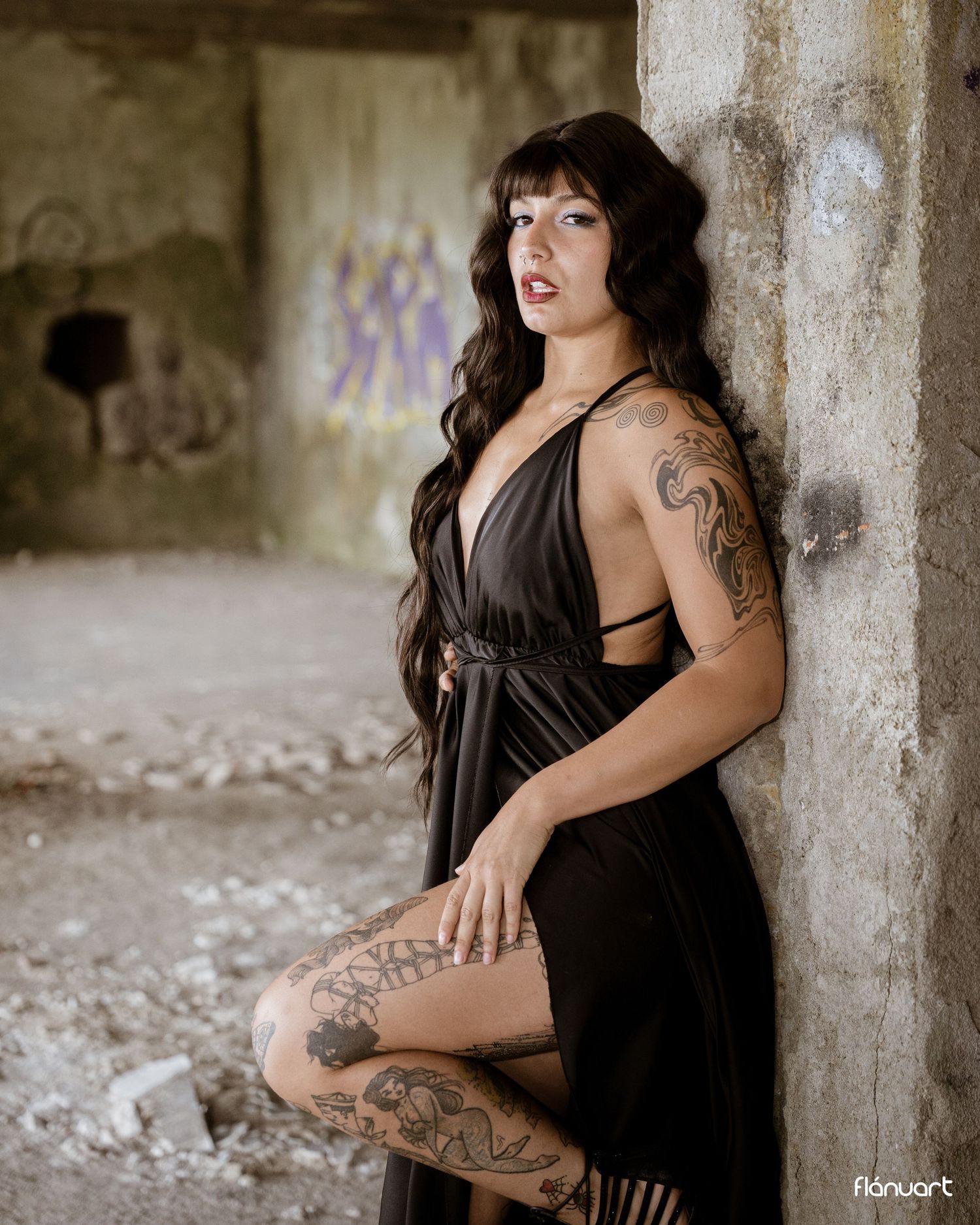 Tattooed woman in black dress posing against concrete pillar in abandoned building with graffiti.