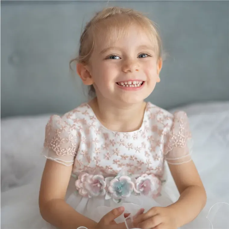 Adorable smiling young girl wearing a white floral embroidered dress with pastel flower accents.