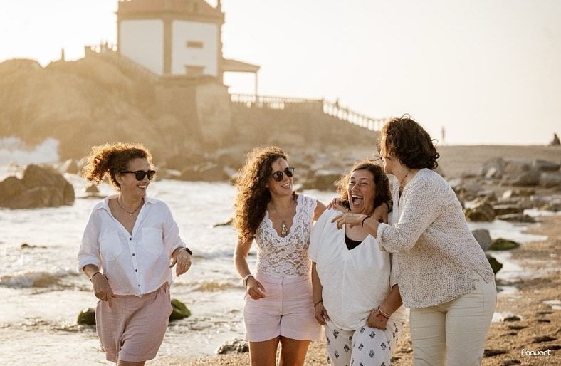 Four women laughing and walking together on a sunny beach near a lighthouse at golden hour.