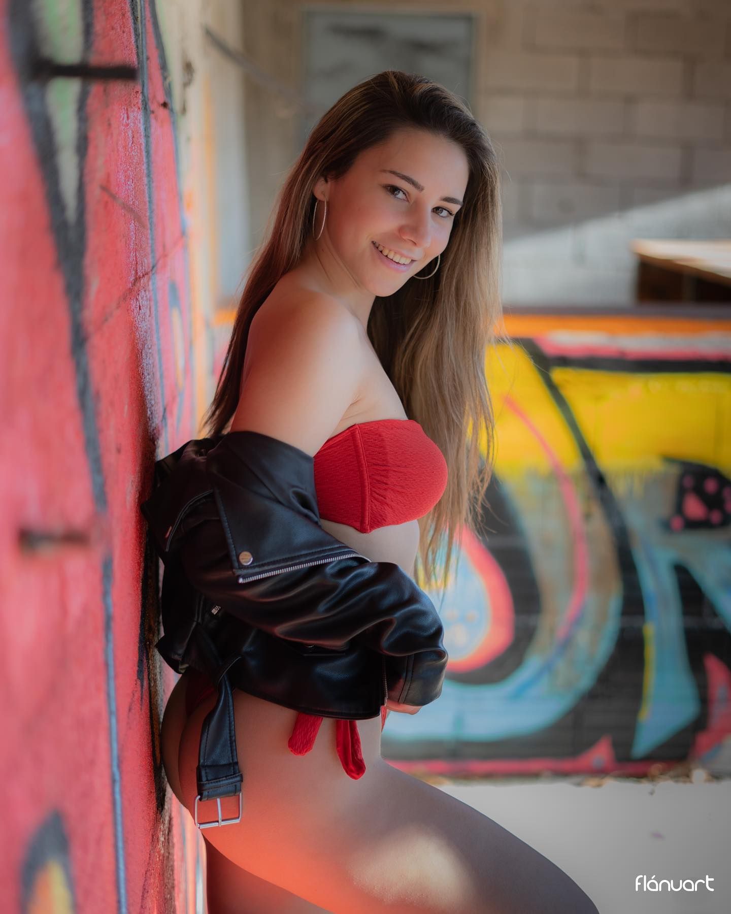 Woman in red strapless top and leather jacket posing by colorful graffiti wall.