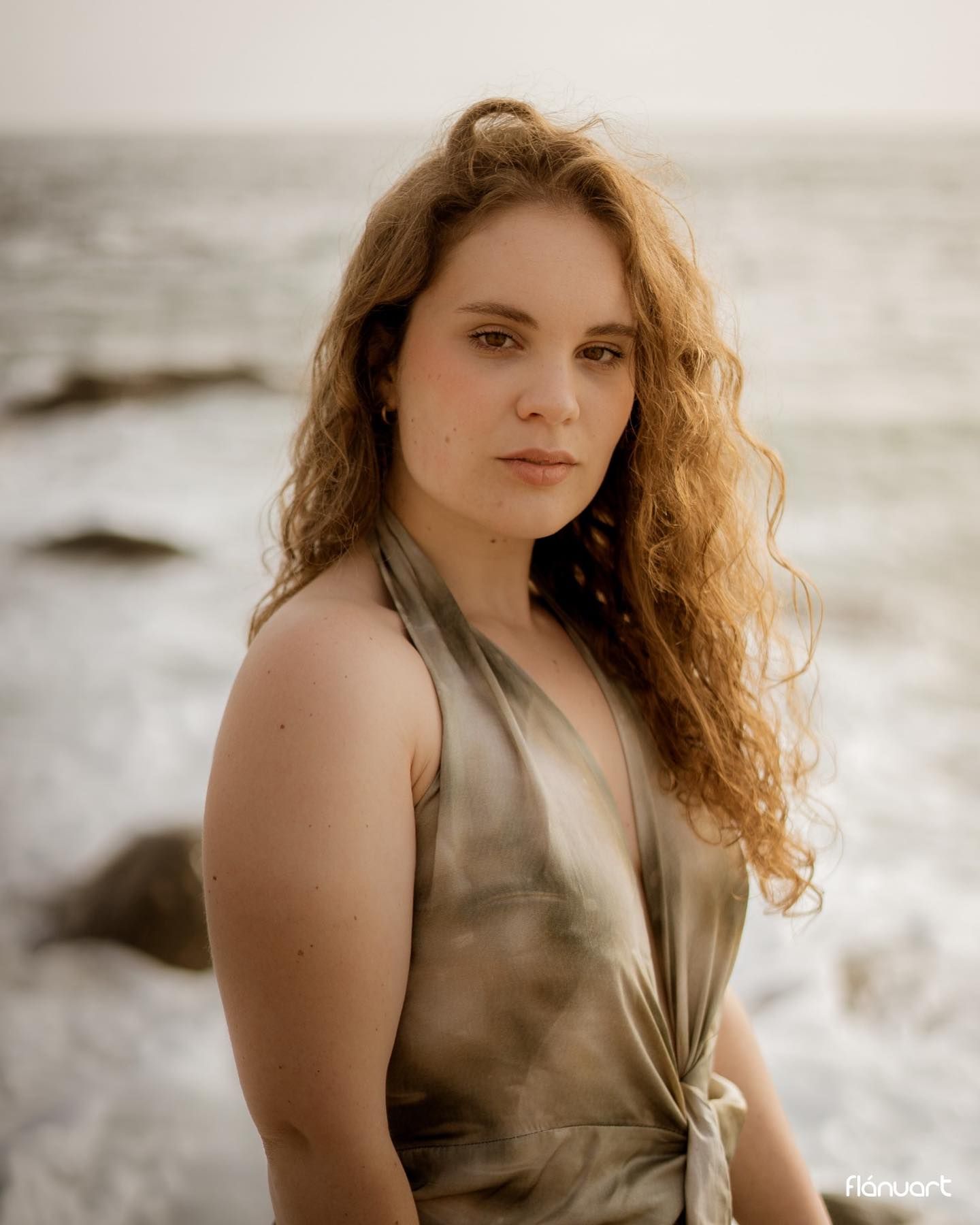 Woman with curly auburn hair in olive wrap dress sitting near ocean with soft evening light.
