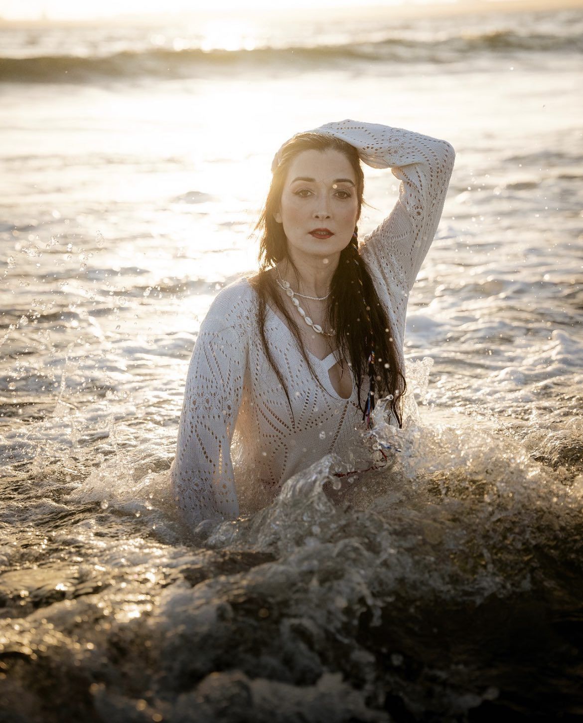Woman in wet white knit sweater sitting in ocean waves at golden sunset on beach.