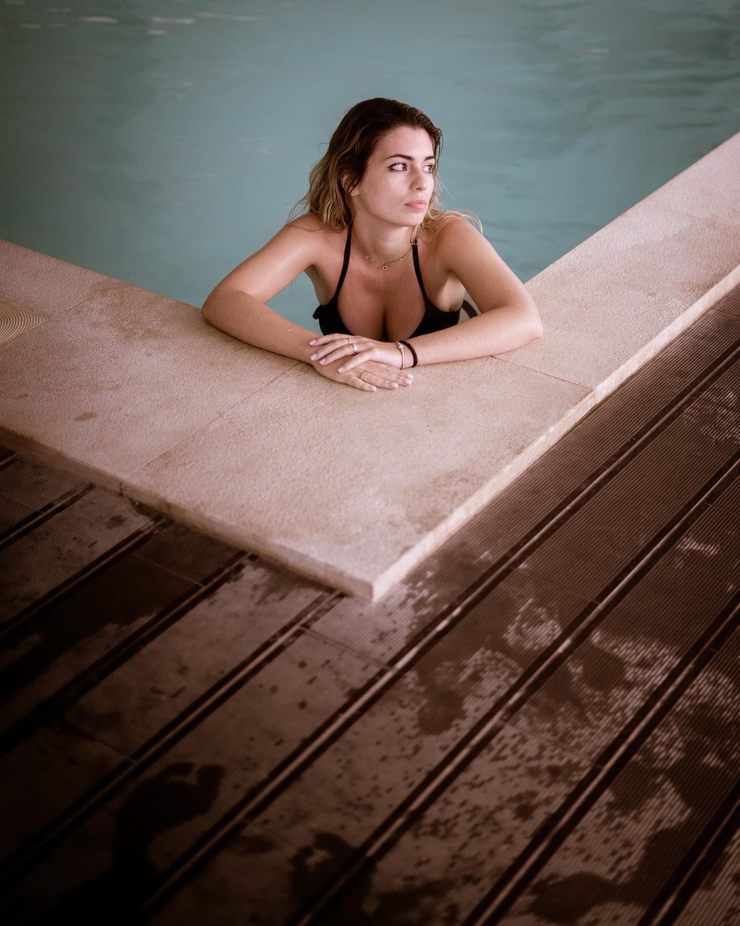 Woman in black bikini resting arms on pool edge, gazing thoughtfully in moody poolside setting.
