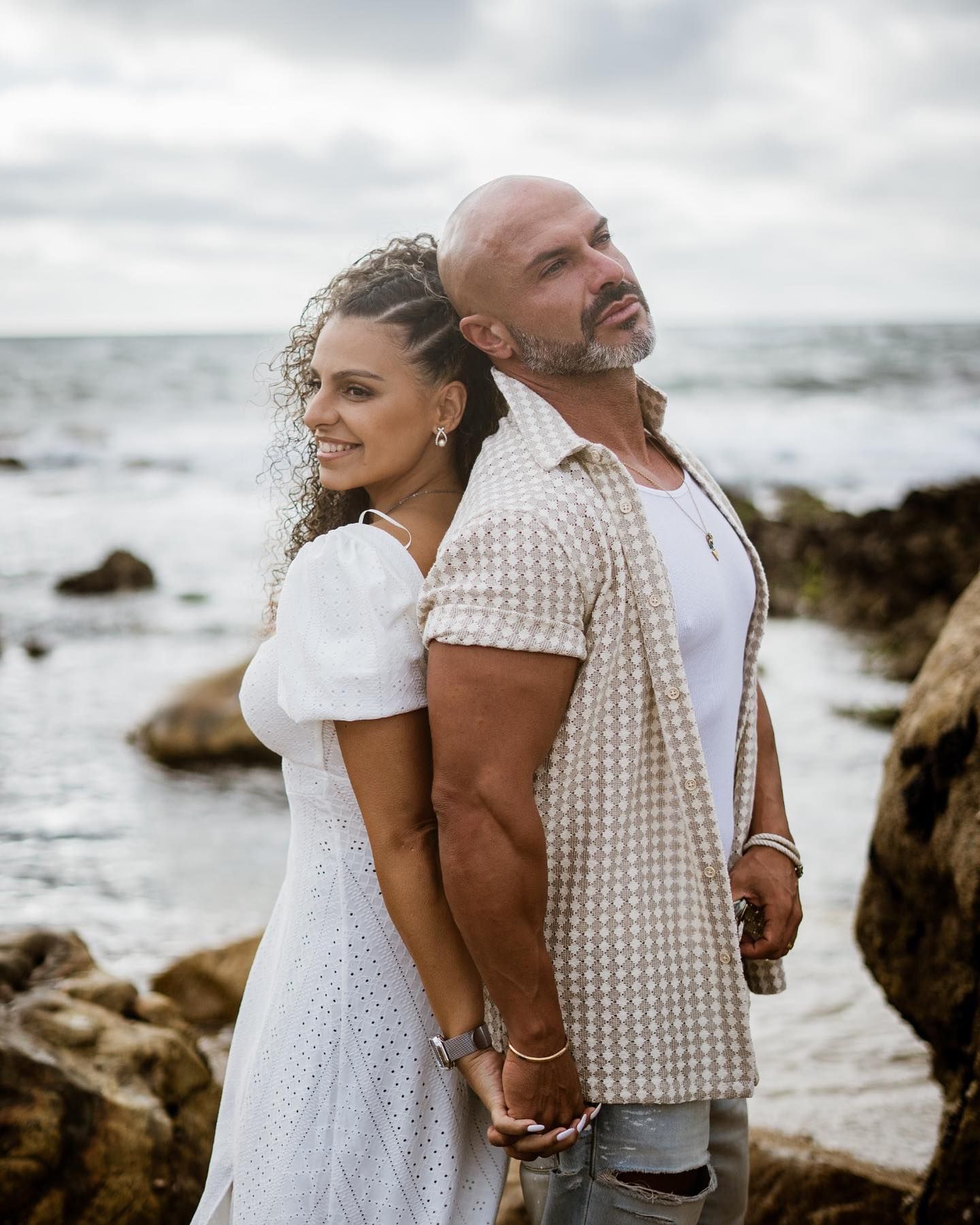 Smiling couple standing back-to-back on rocky beach, woman in white dress holding hands with bald man.