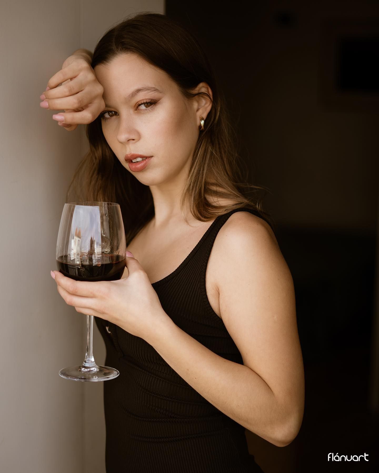 Young woman in black dress holding a glass of red wine leaning against a wall.