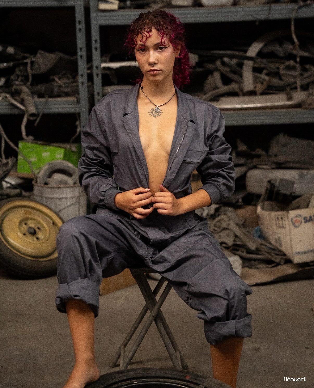 Woman with red curly hair sits on a stool in an open gray jumpsuit in an auto repair garage.