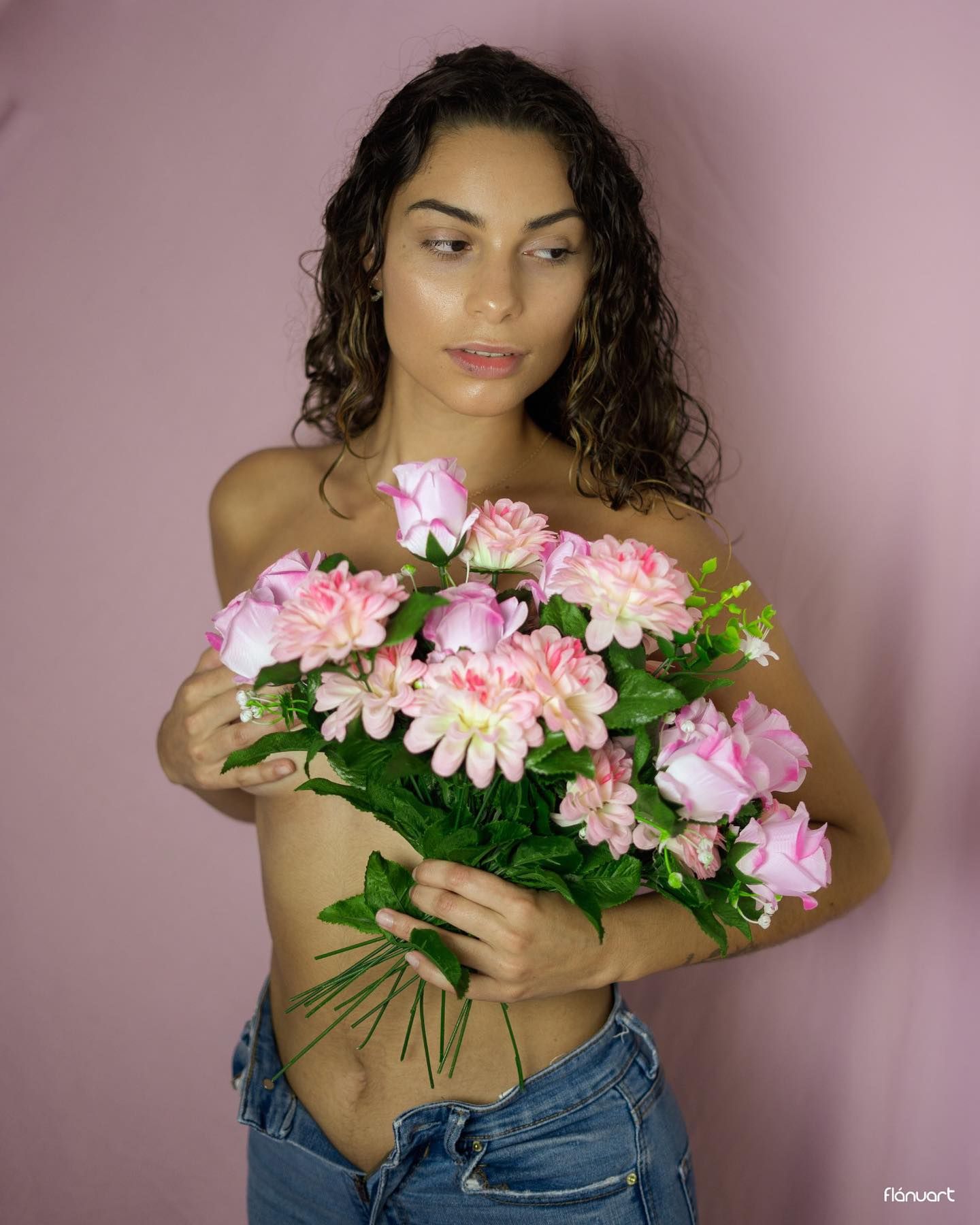 Curly-haired woman holding pink flower bouquet topless in jeans against pink background.