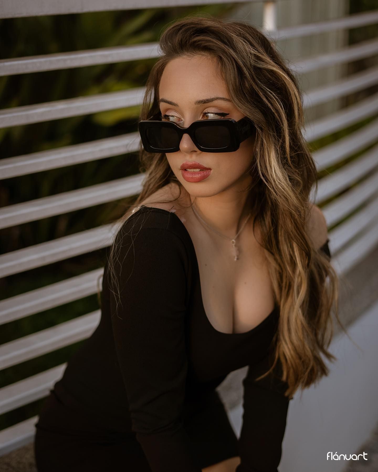 Young woman in black outfit and rectangular sunglasses posing outdoors with long highlighted hair.