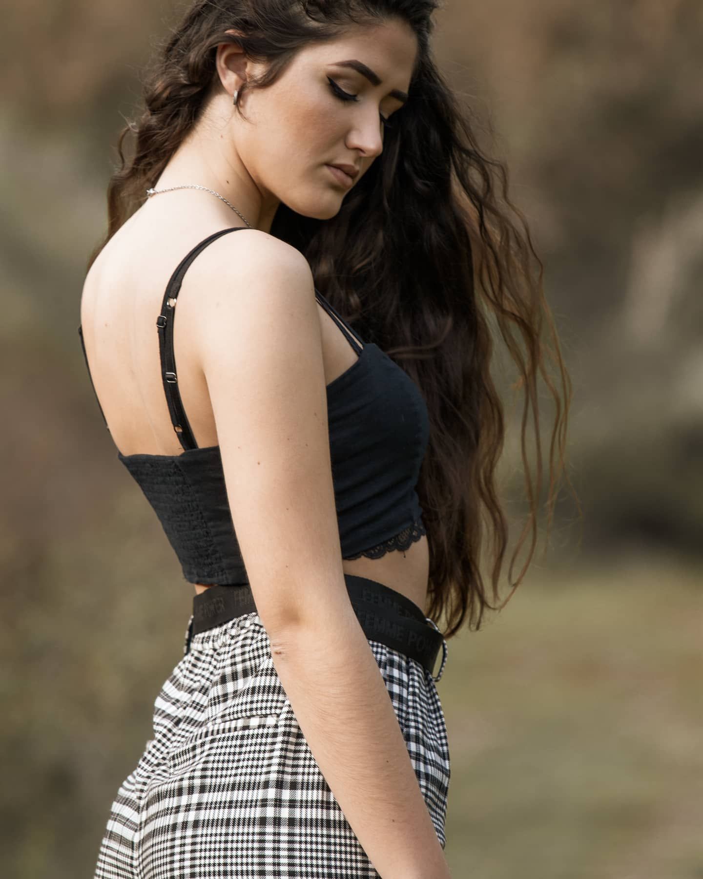 Young woman in black crop top and plaid pants poses with back to camera outdoors.