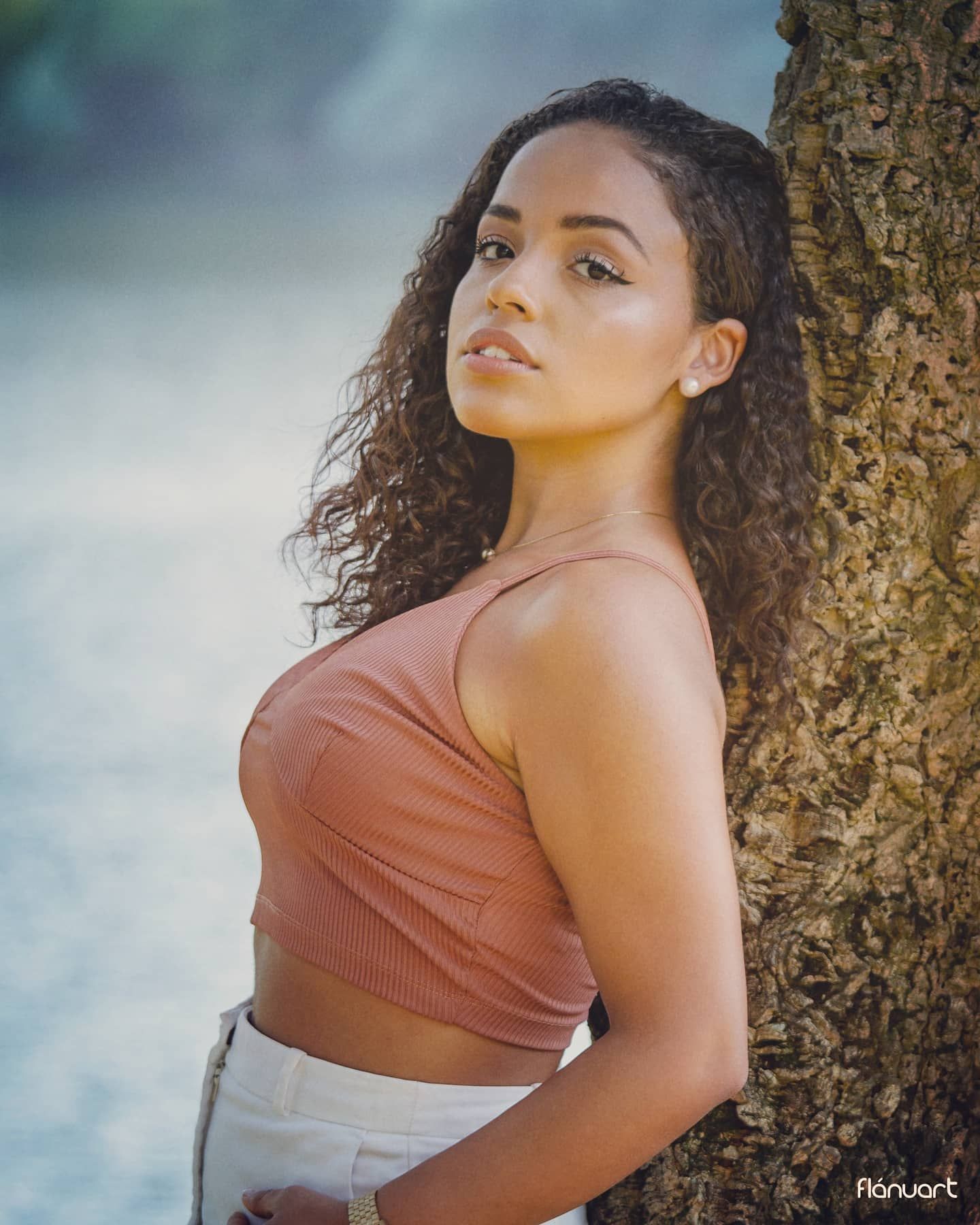 Young woman with curly hair in a pink crop top leaning against a tree outdoors.