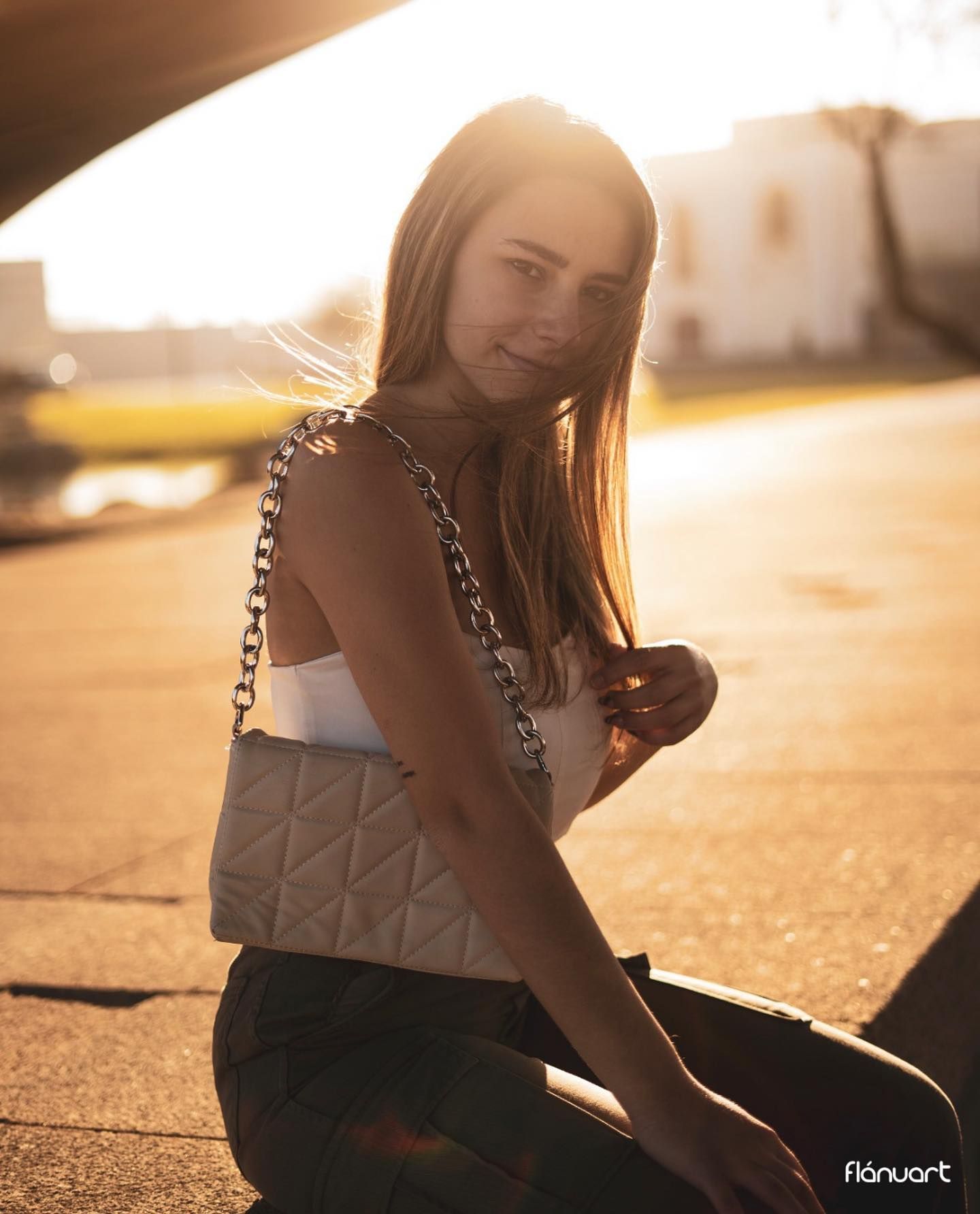 Young woman with white quilted shoulder bag sitting outdoors in golden sunset light.