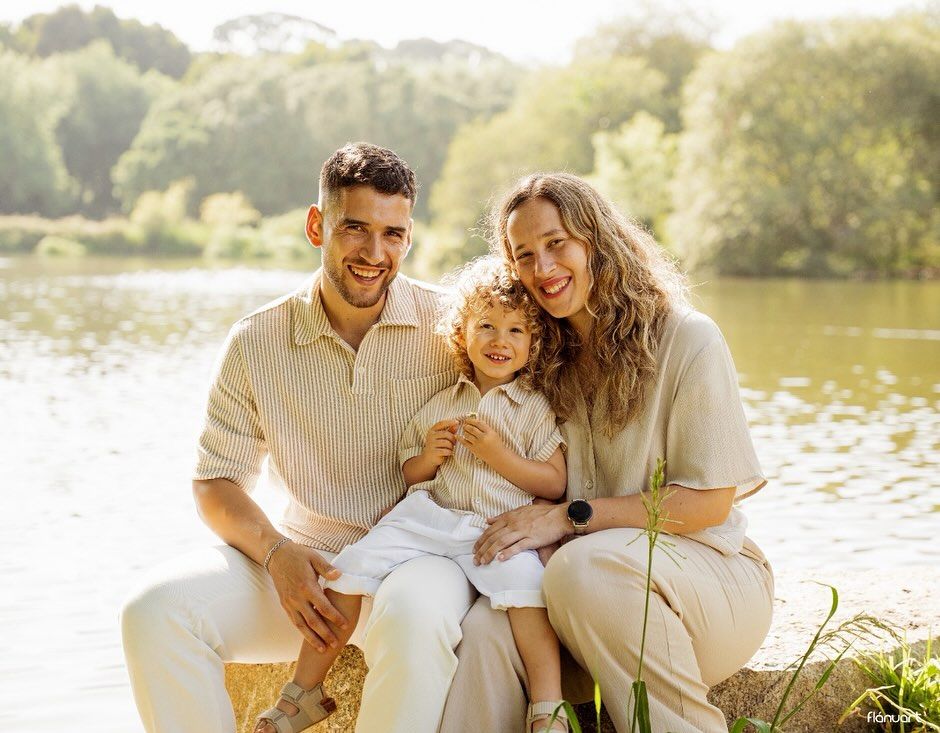 Happy family of three posing by a serene lake, parents smiling with their curly-haired toddler on a sunny day.