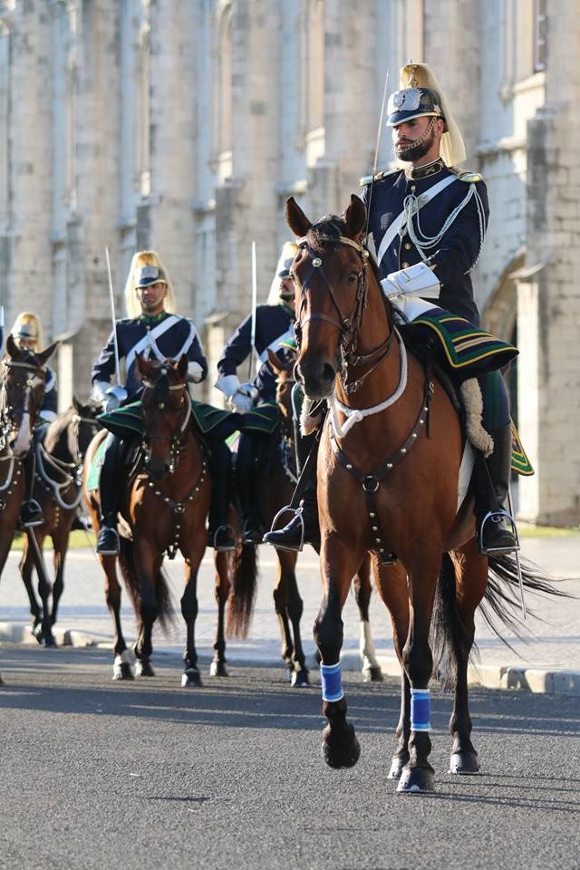 Military cavalry officers in formal uniforms riding brown horses in a ceremonial parade.