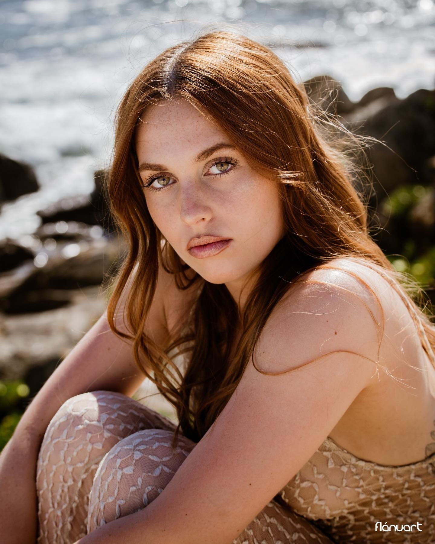 Young redhead woman in lace outfit sitting by rocky oceanside in warm natural light.