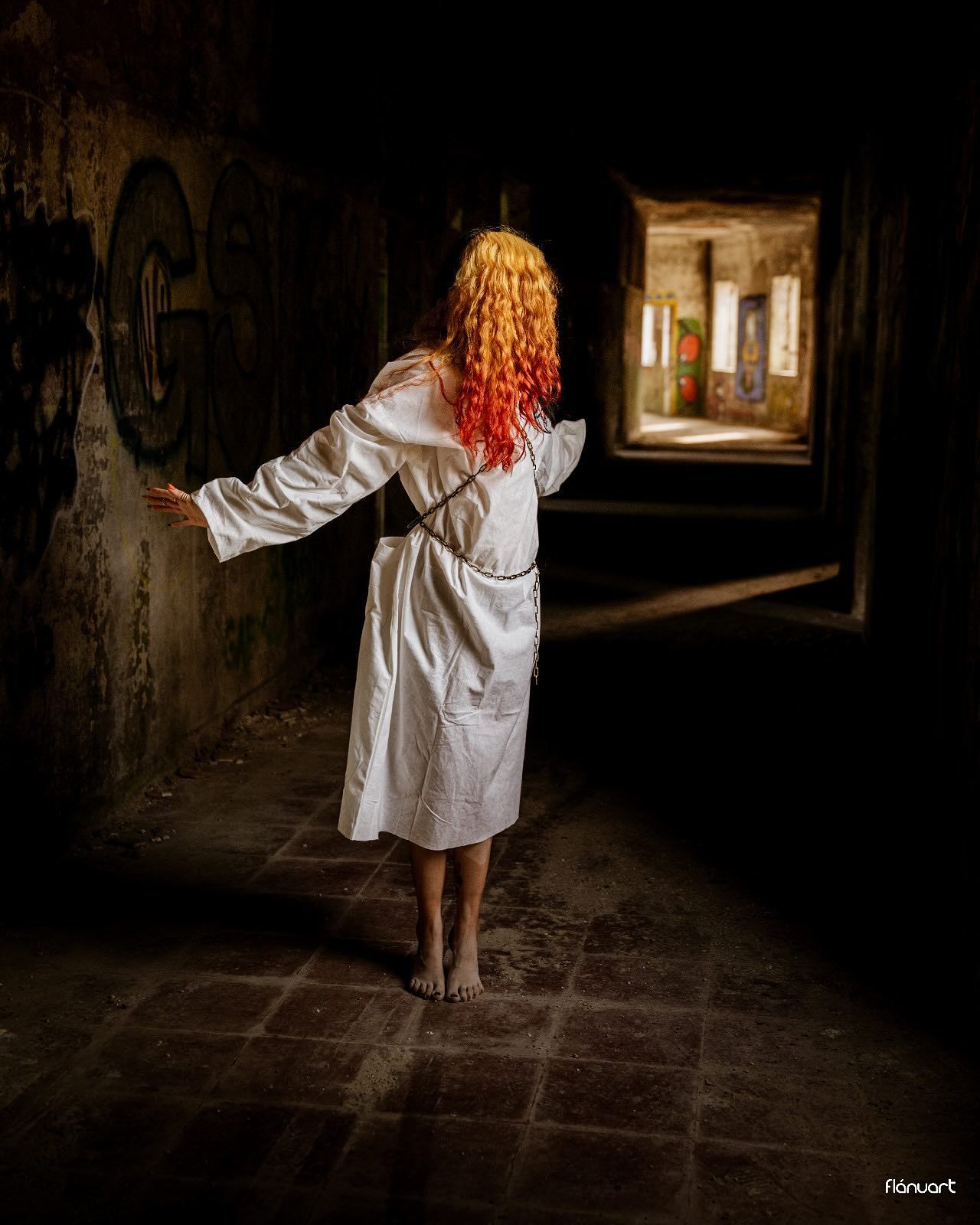 Woman with vibrant red curly hair in white dress standing in dark abandoned building hallway.