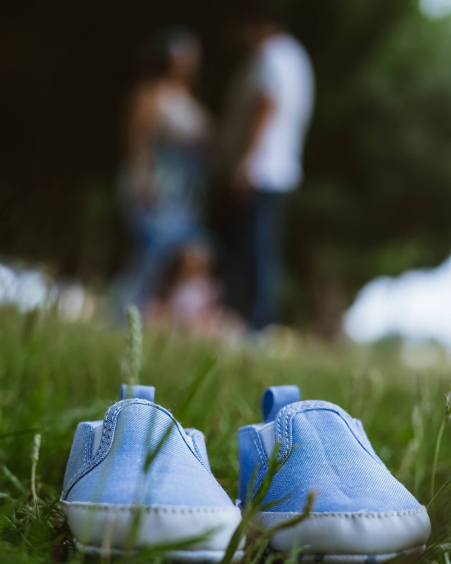 Tiny blue baby shoes in grass with couple standing blurred in background, pregnancy announcement.