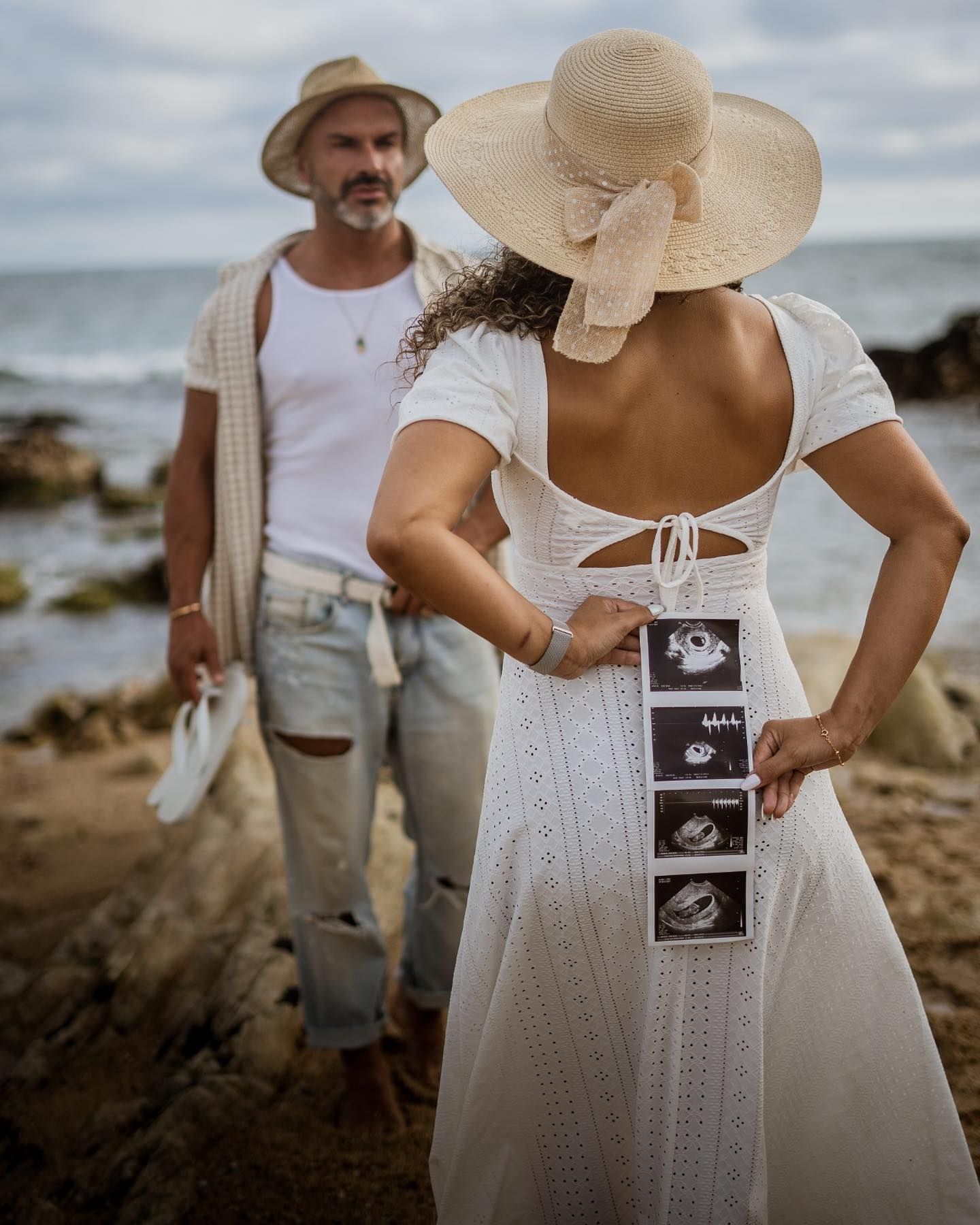 A pregnant woman in a white dress holds ultrasound photos behind her back near a rocky beach.