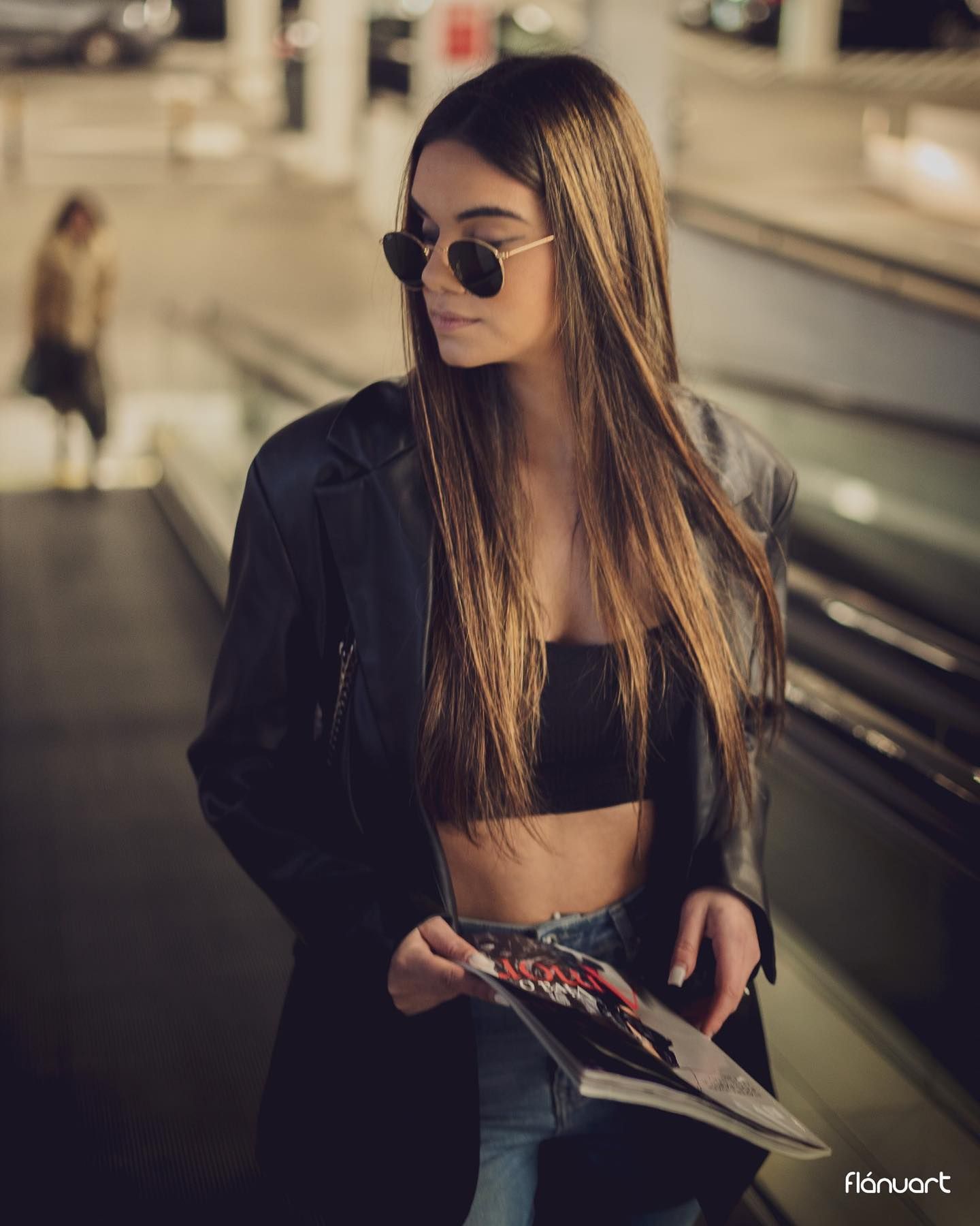 Woman in black crop top and blazer holds magazine wearing round sunglasses at airport.