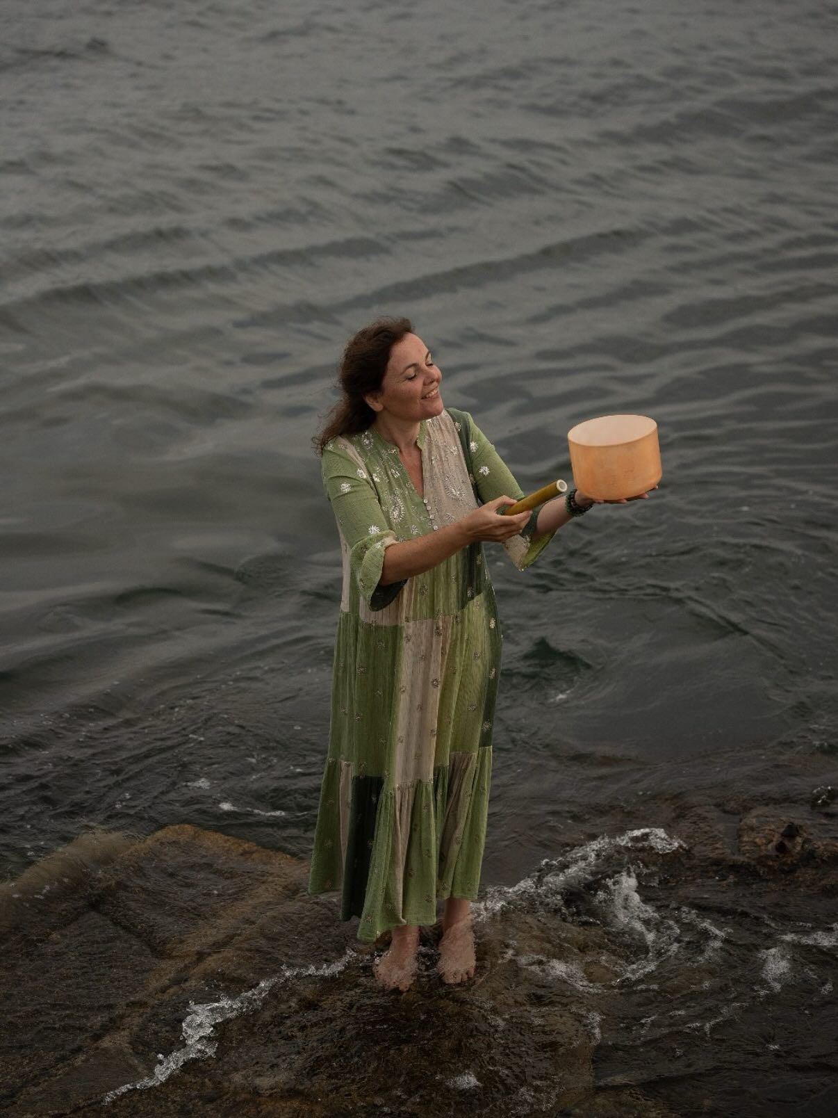 Woman in green floral dress standing on rocks by the ocean holding a wooden bowl.